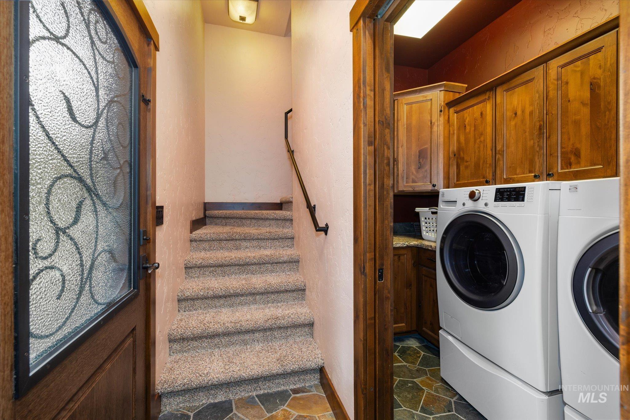 Entryway featuring a decorative glass-paneled door with dark wood framing, carpeted stairs with a dark wood handrail, and a laundry area with wood cabinetry, a countertop, and flagstone flooring