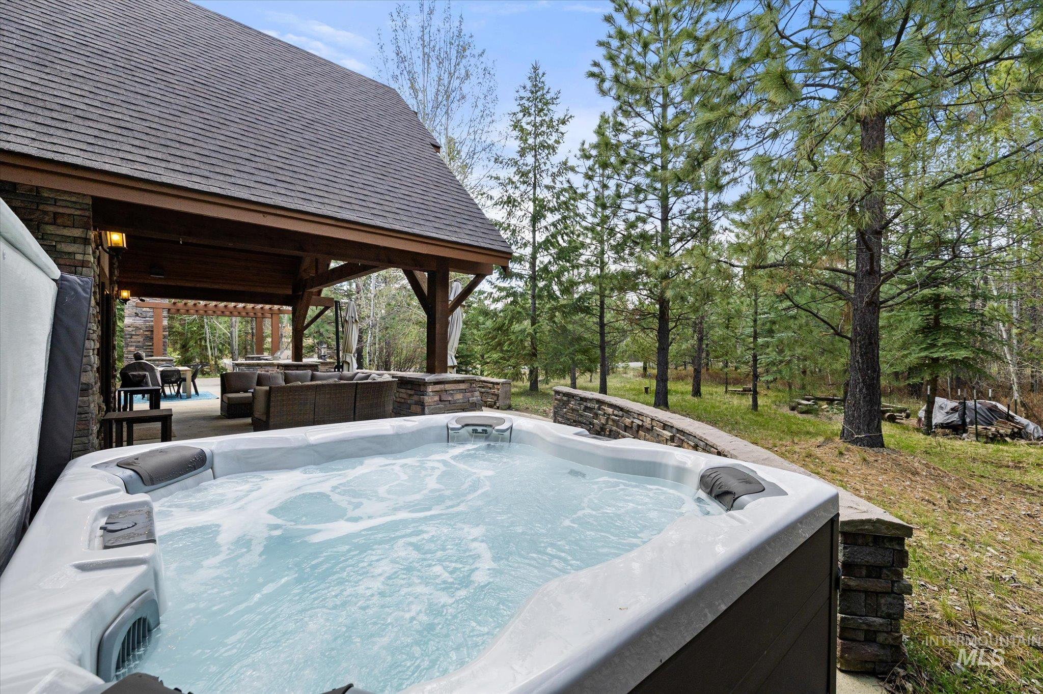 Outdoor spa with bubbling water, stone retaining wall, and a covered patio area with exposed timber beams and an asphalt shingle roof
