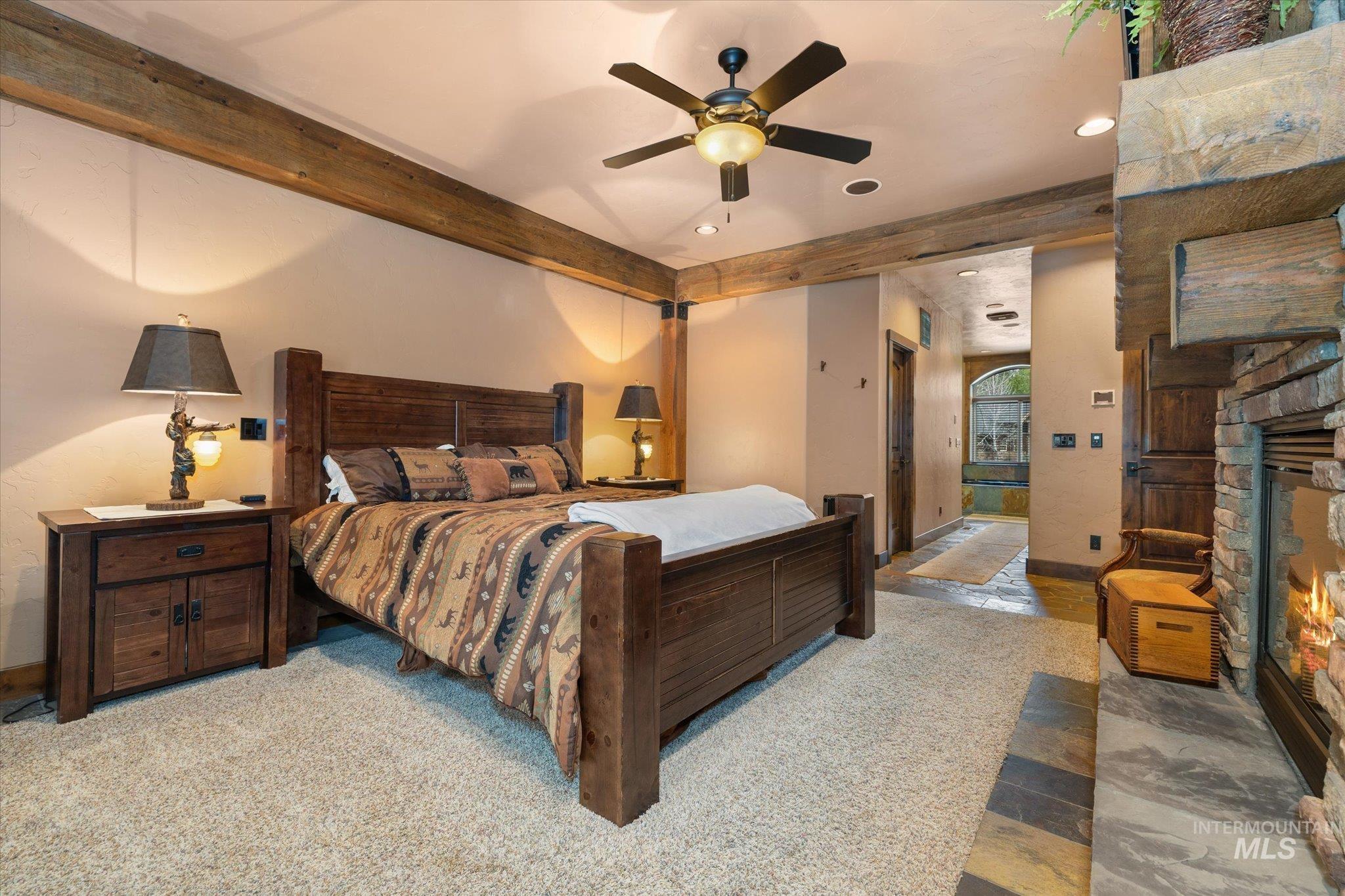 Carpeted bedroom featuring exposed wood beams, a ceiling fan, a stone fireplace, and an adjacent stone-tiled hallway