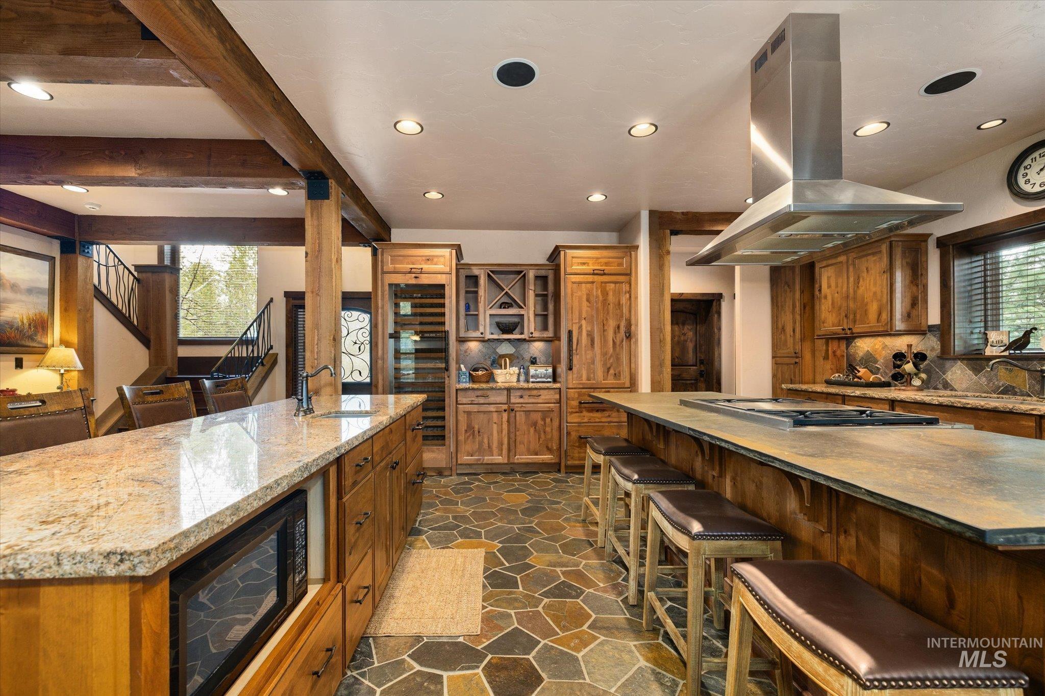 Spacious kitchen featuring two large islands with stone countertops, a built-in cooktop with a stainless steel range hood, stone tile flooring, and exposed wood beams
