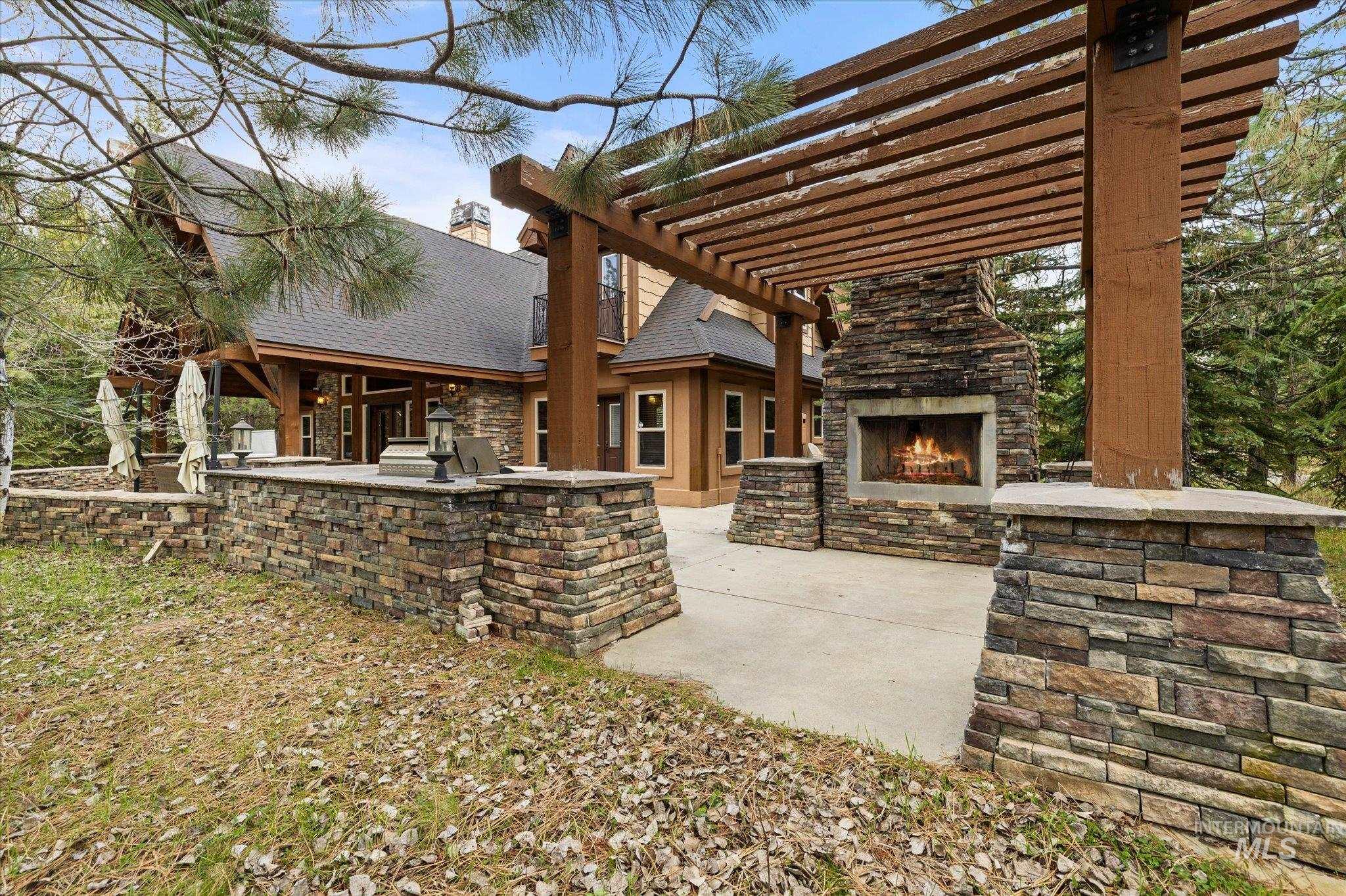 Outdoor patio featuring a stone fireplace under a wood pergola, integrated stone planters, a paved surface, and exterior lighting fixtures
