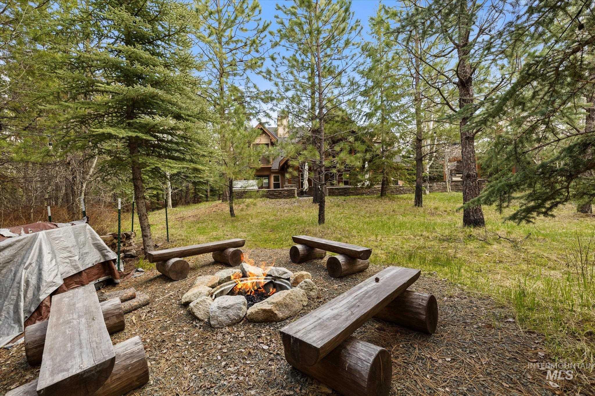 Outdoor fire pit with natural stone surround and log benches, set within a wooded landscape featuring mature trees and a distant dwelling