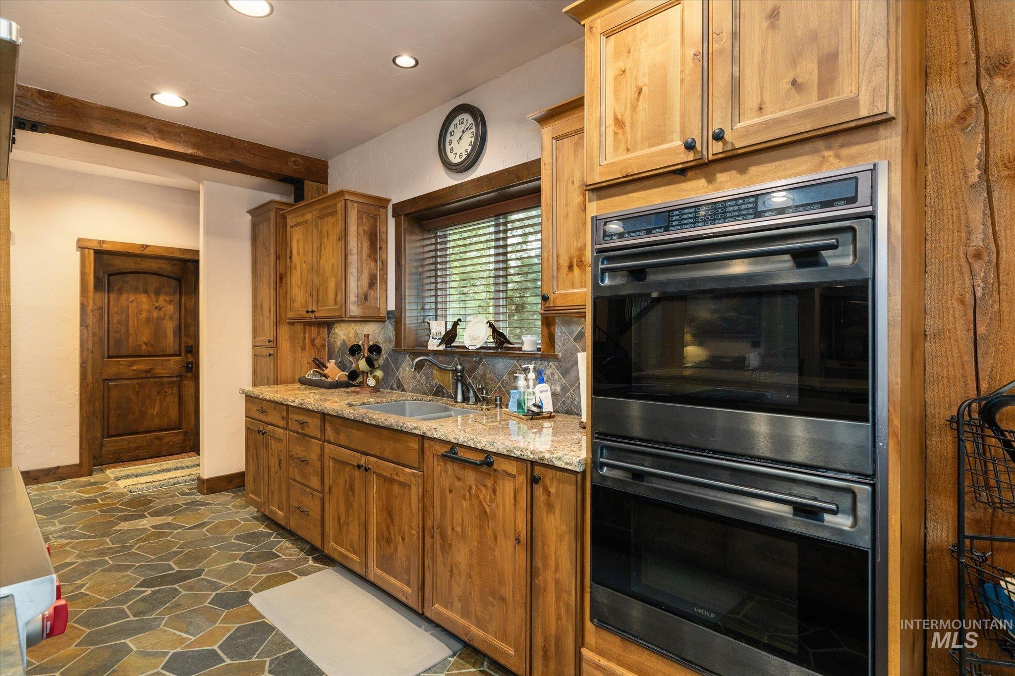 Kitchen featuring stacked stainless steel ovens, wood cabinetry, granite countertops, and stone tile flooring