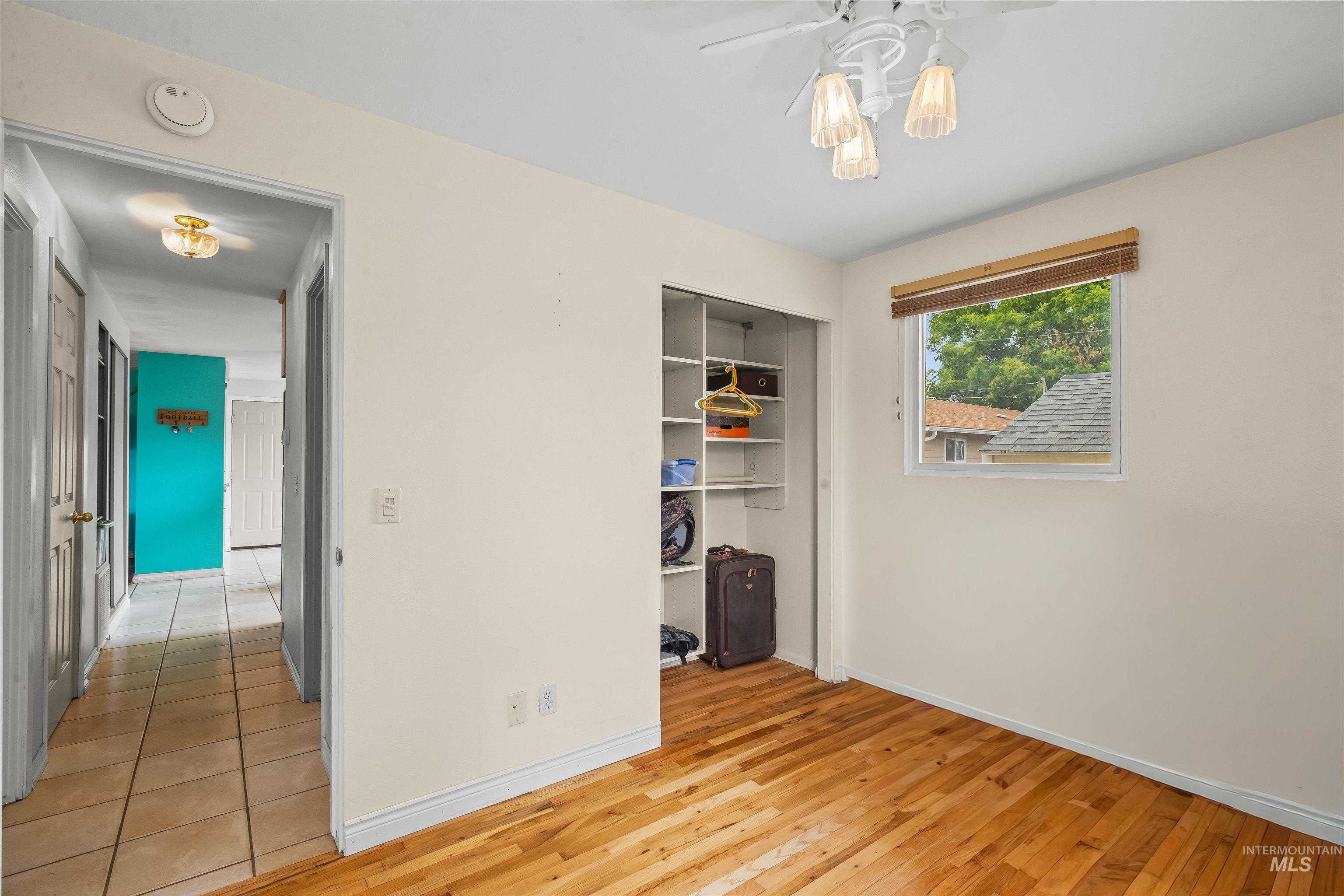 Room featuring wood-finish flooring, a window with a natural wood-tone blind, a ceiling fan with integrated lighting, and a built-in closet with shelving