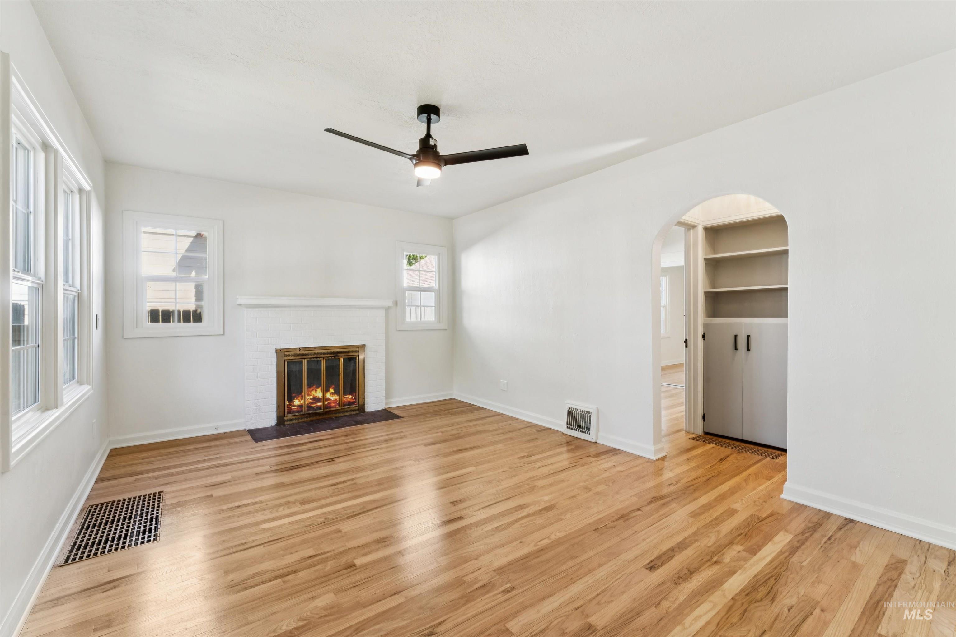Unfurnished living room with light wood-style floors, a brick fireplace, arched walkways, and a ceiling fan