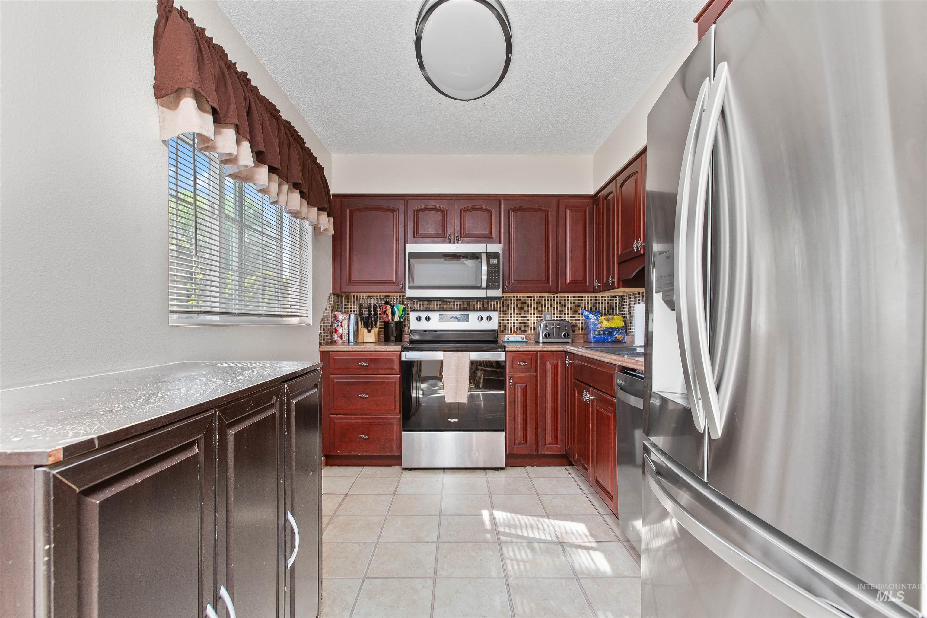 Kitchen with stainless steel appliances, a textured ceiling, light tile patterned flooring, decorative backsplash, and reddish brown cabinets