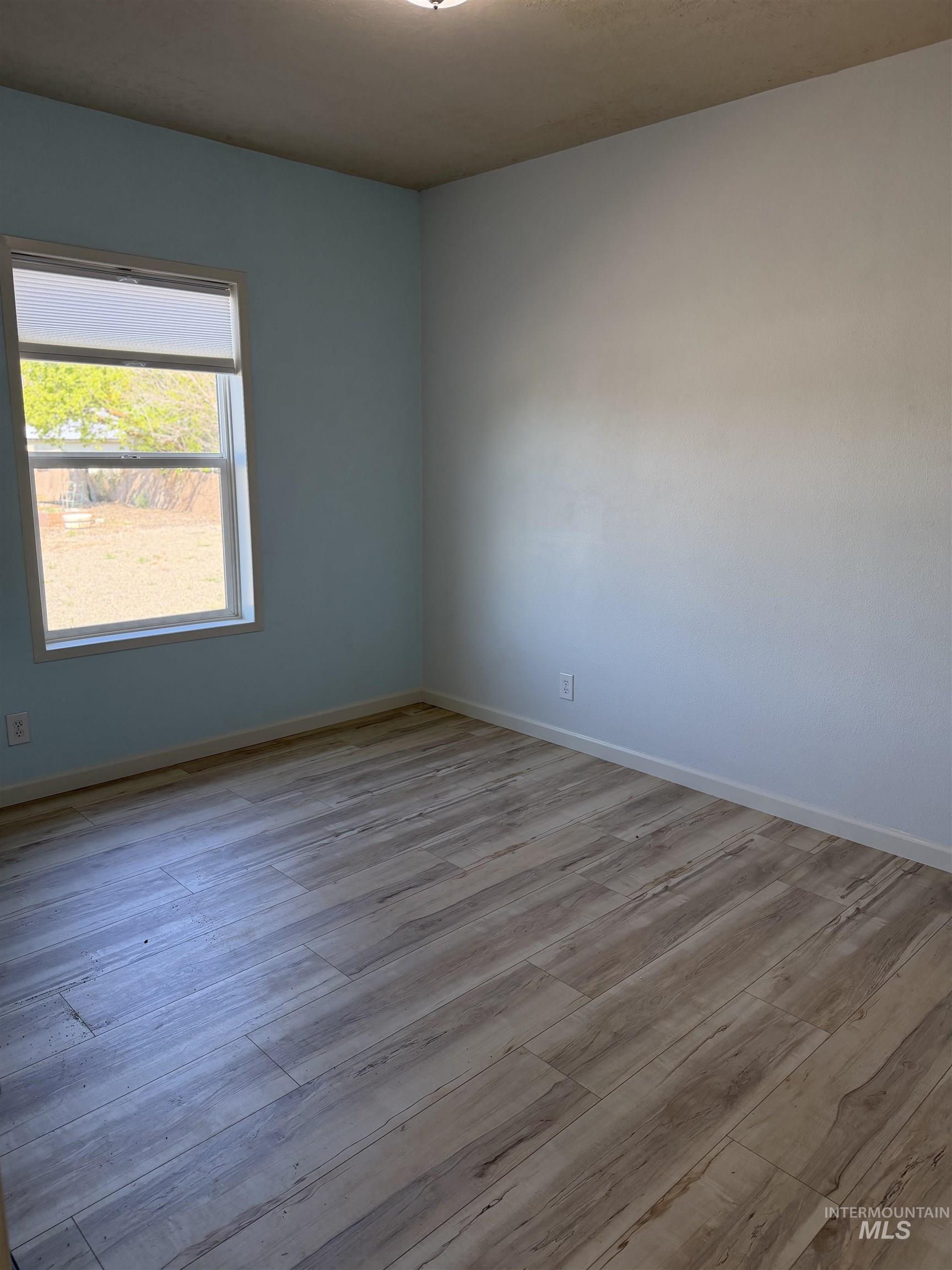 Spacious room featuring light wood-finish flooring, a single window with blinds, and a flush-mount ceiling light