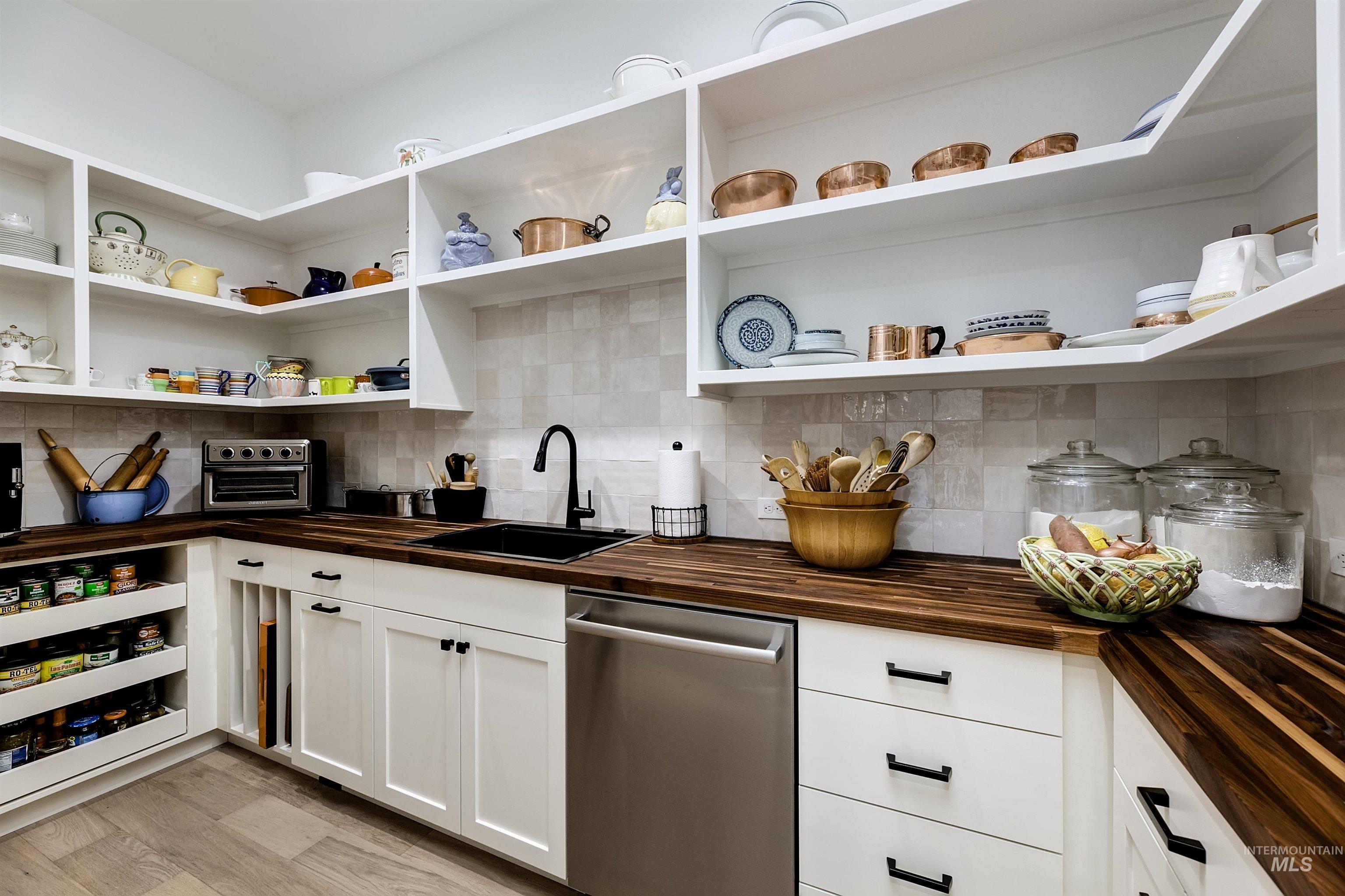 Corner pantry featuring extensive open shelving, white cabinetry, dark wood-finish countertops, a black undermount sink, and a stainless steel dishwasher