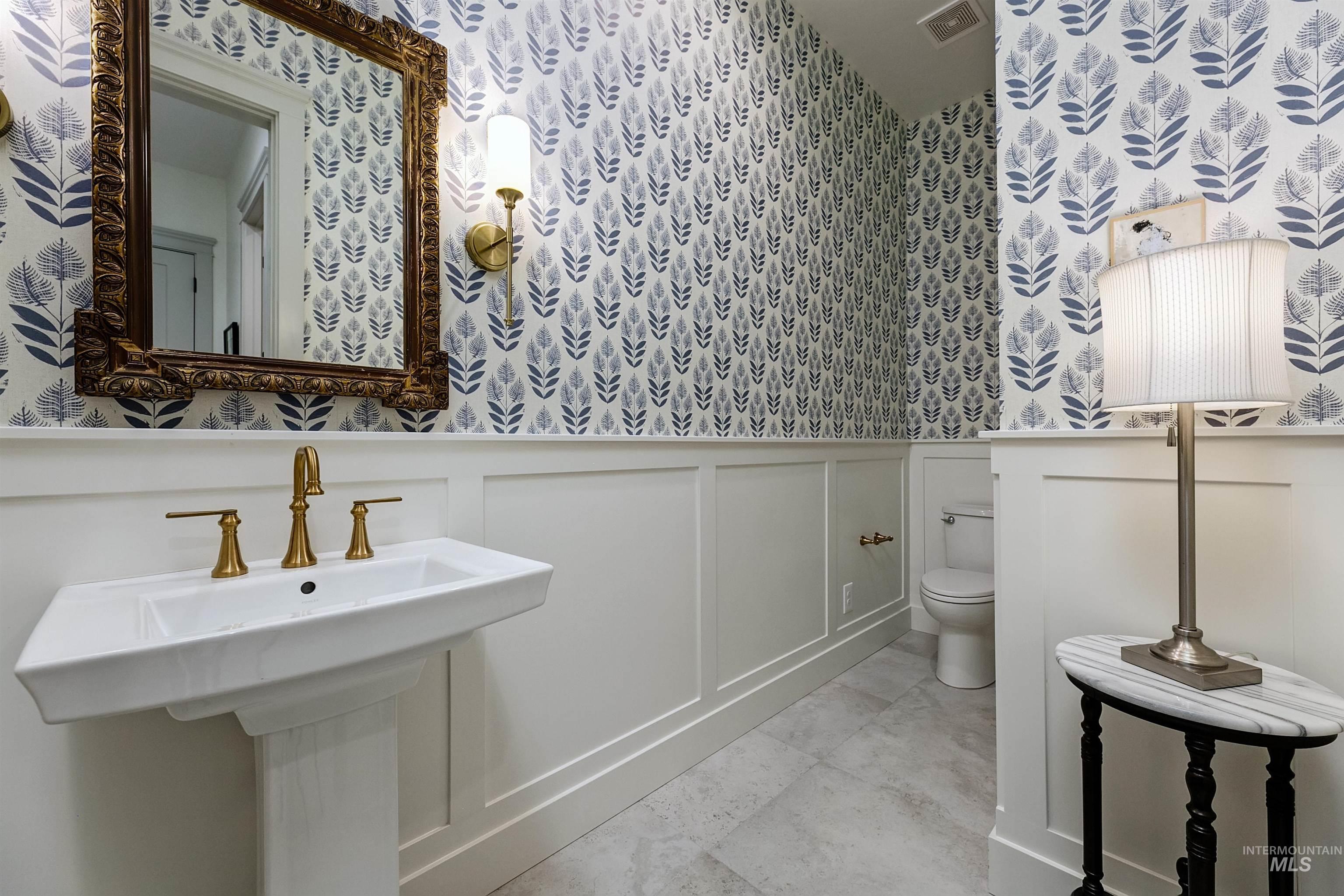 Pedestal sink with brass-finish faucet, framed mirror, wall sconce lighting, wainscoting, and patterned wallpaper