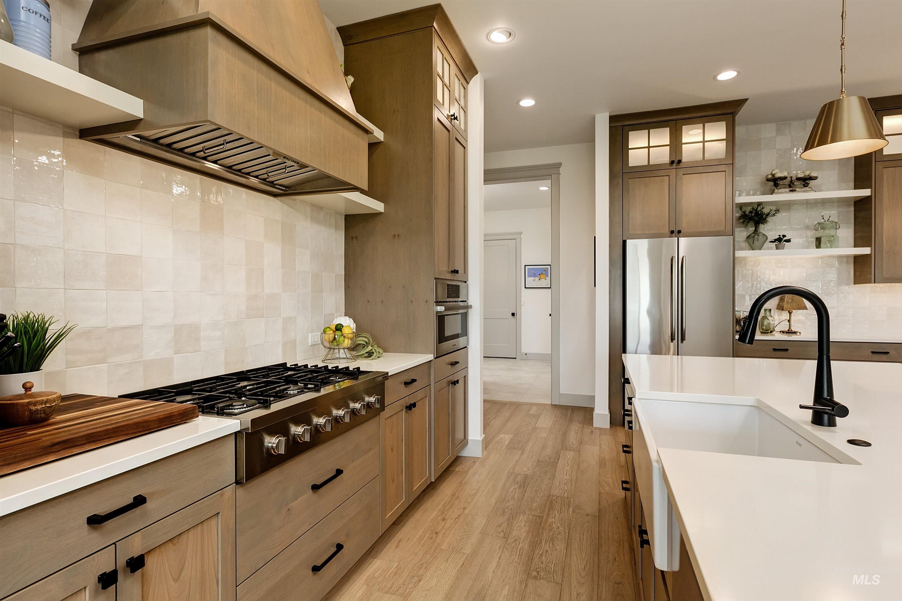 Gourmet kitchen featuring a built-in gas range, coordinating wood-finish range hood, light-toned tile backsplash, light wood-finish cabinetry with black hardware, and an undermount sink with a black gooseneck faucet
