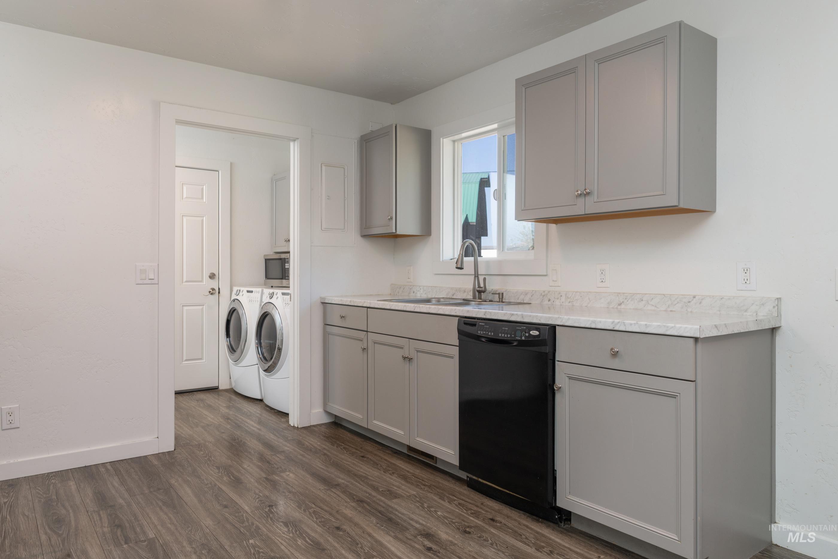 Kitchen with gray cabinetry, washing machine and clothes dryer, light countertops, dark wood-type flooring, and black dishwasher