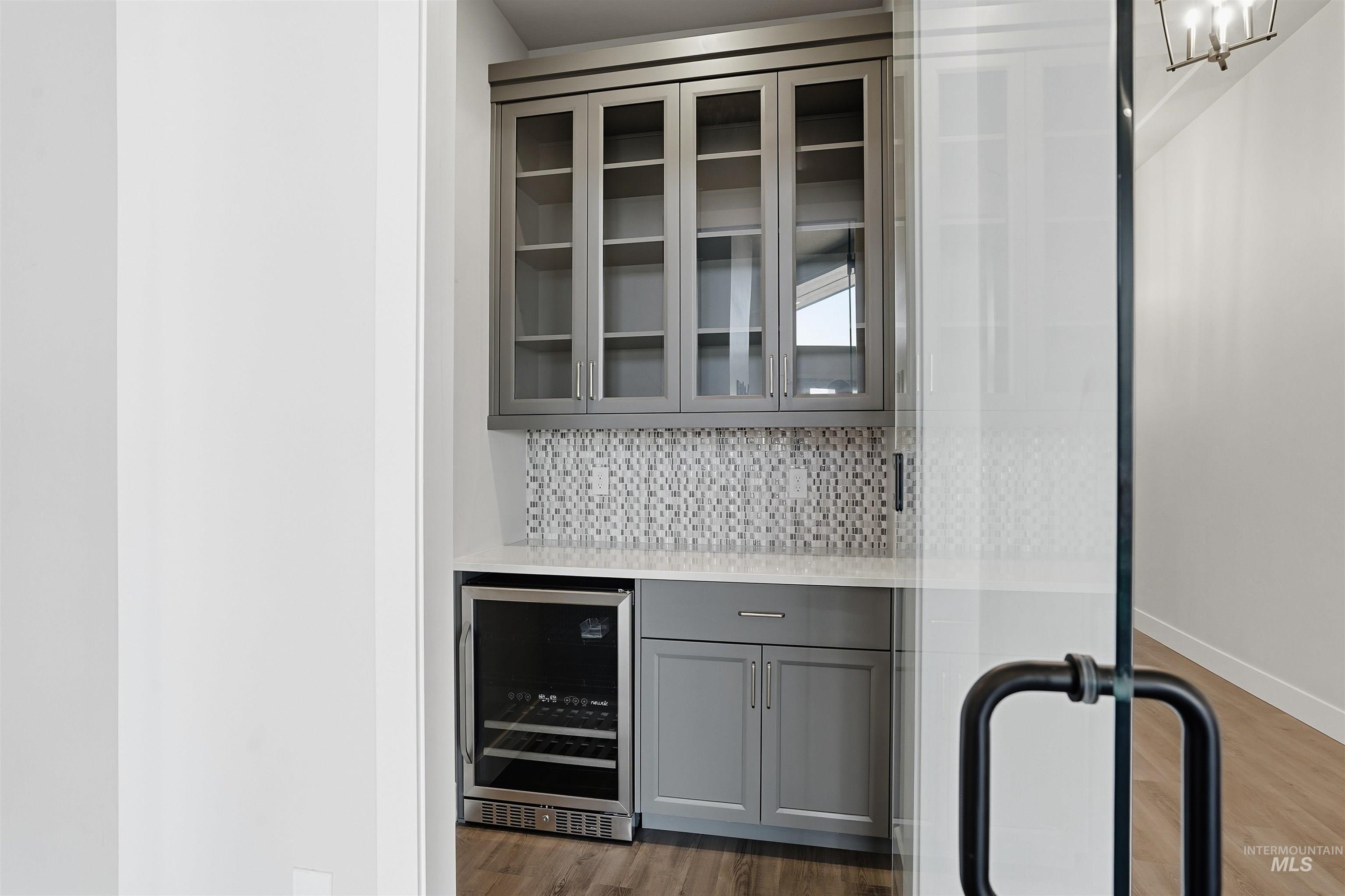 Indoor dry bar featuring gray cabinetry, beverage cooler, decorative backsplash, light stone counters, and glass insert cabinets
