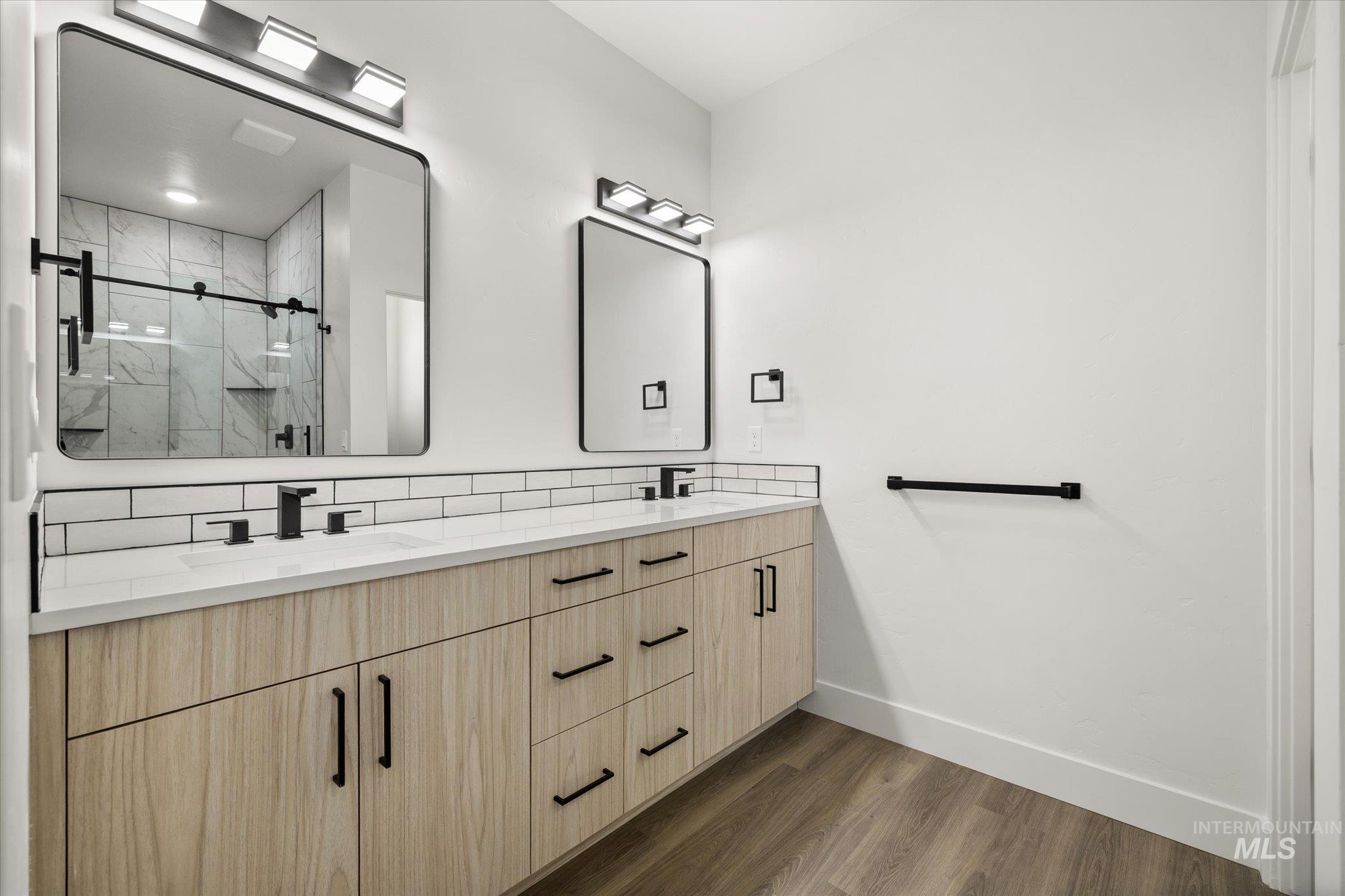 Bathroom featuring dark wood-style flooring, double vanity, a marble finish shower, and backsplash