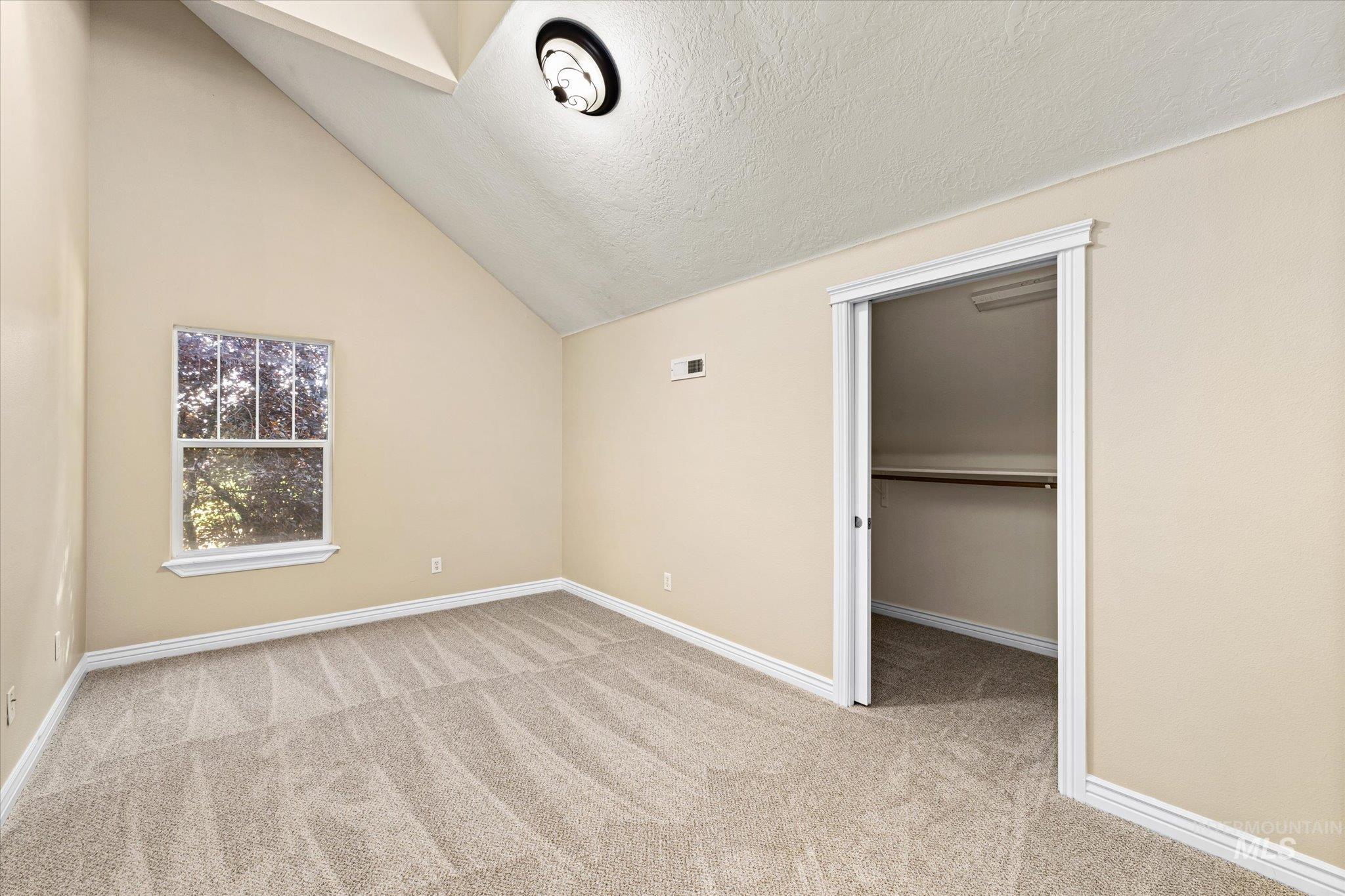 Unfurnished bedroom with a walk in closet, a textured ceiling, lofted ceiling, and light colored carpet