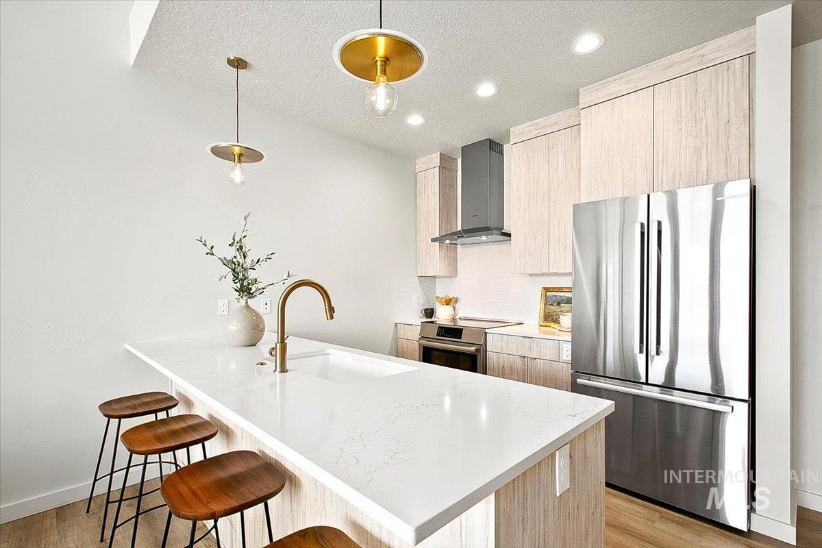 Kitchen featuring light brown cabinetry, modern cabinets, stainless steel appliances, wall chimney range hood, and a textured ceiling