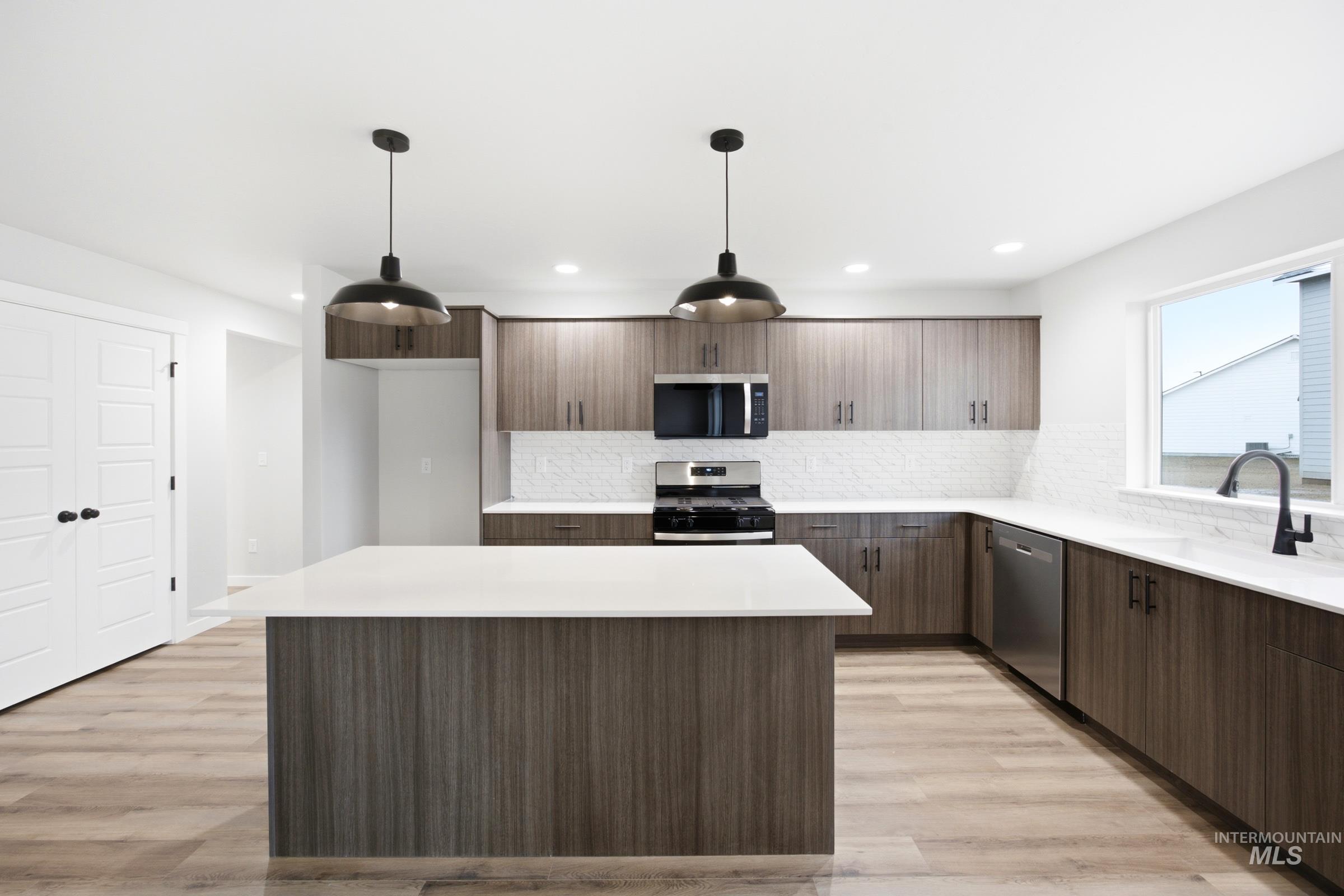 Modern kitchen featuring a central island with a solid surface countertop, wood-finish cabinetry, stainless steel appliances, a white subway tile backsplash, and two pendant light fixtures