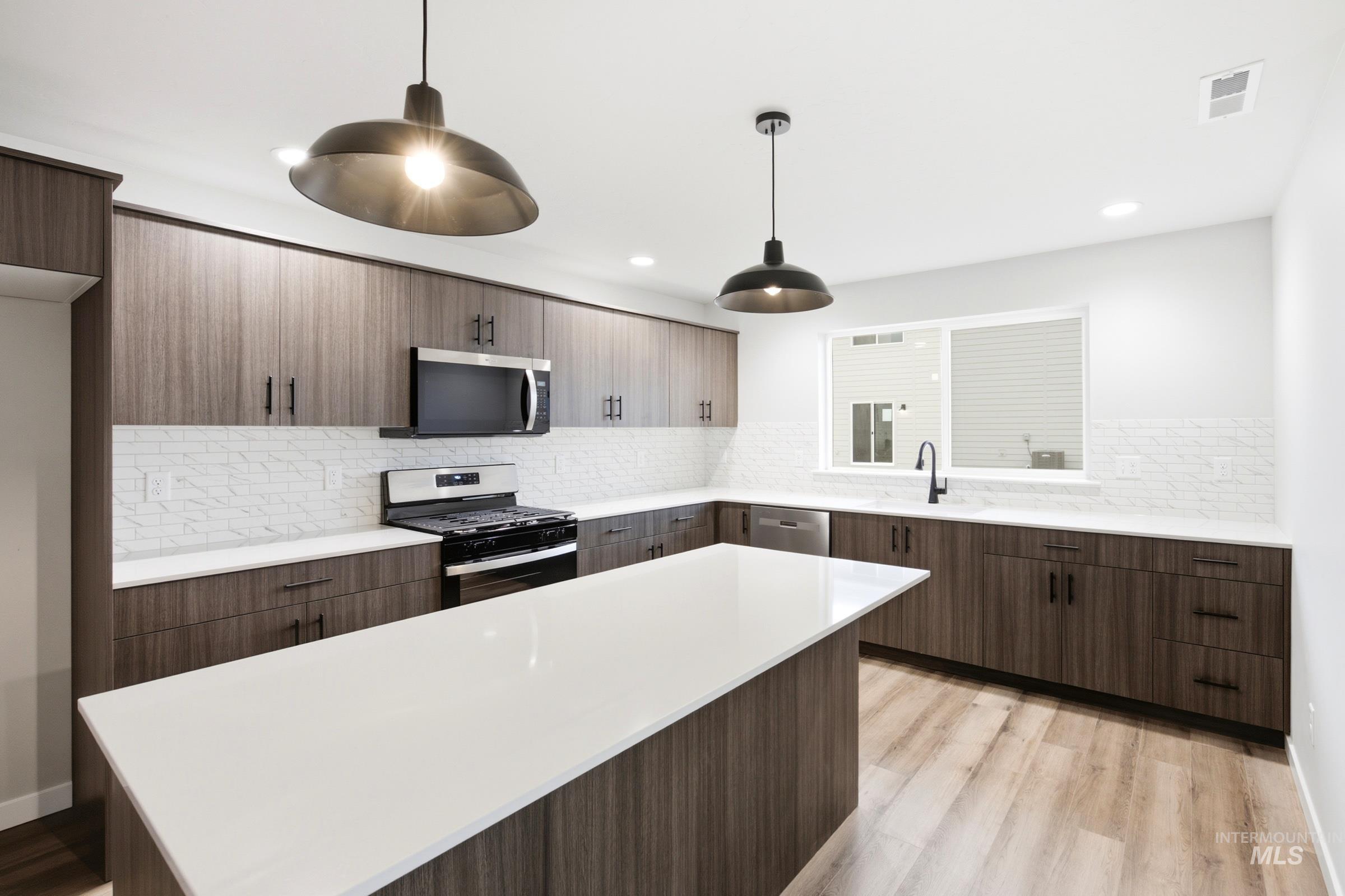 Kitchen featuring an expansive island with a solid surface countertop, wood-finish flooring, dark wood-finish cabinetry, stainless steel appliances, and two industrial-style pendant lights