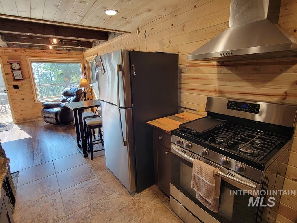 Kitchen with wood walls, appliances with stainless steel finishes, wall chimney exhaust hood, wood ceiling, and light tile patterned floors
