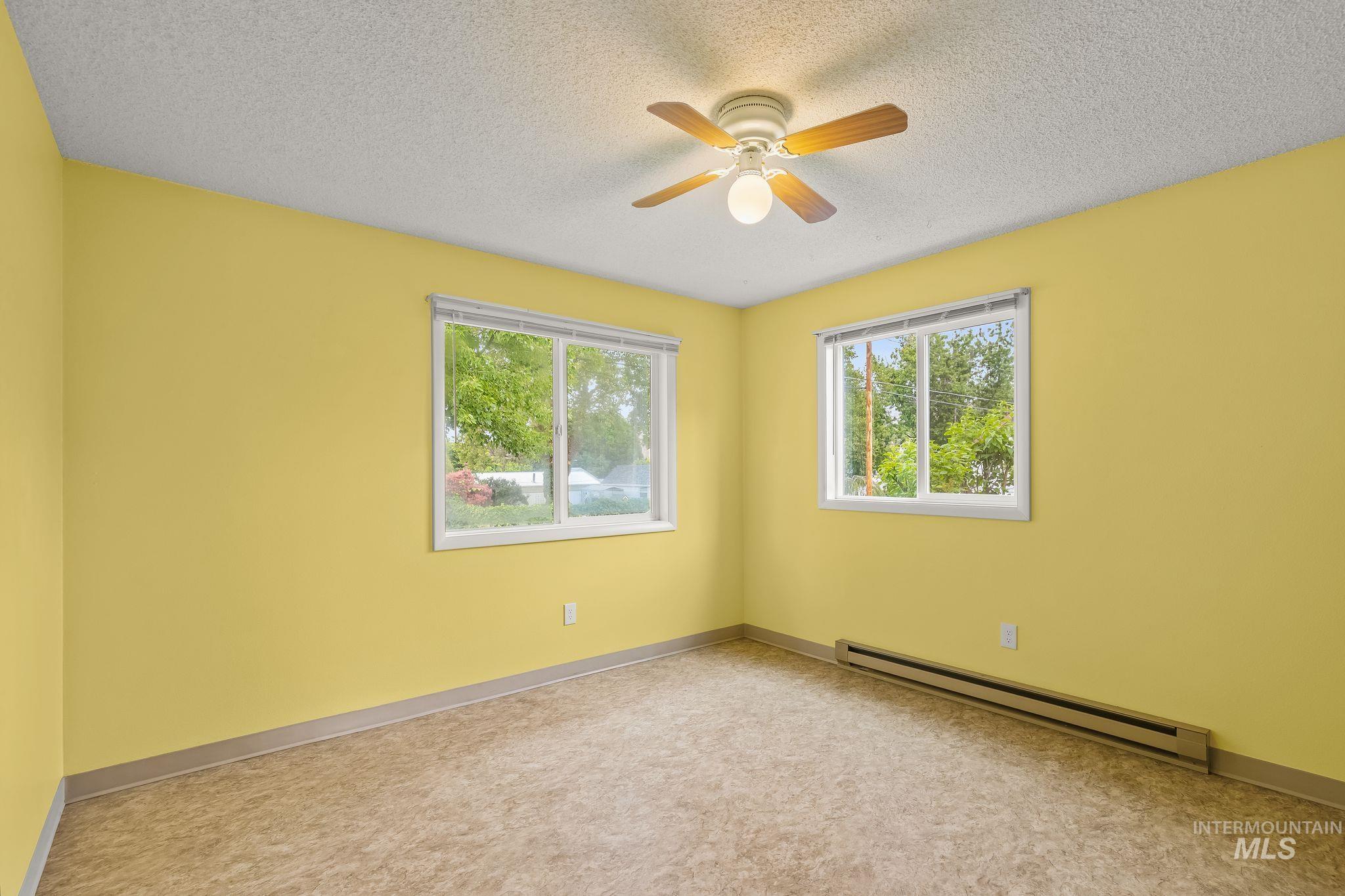 Spacious room featuring two large windows, a ceiling fan with light fixture, and baseboard heating