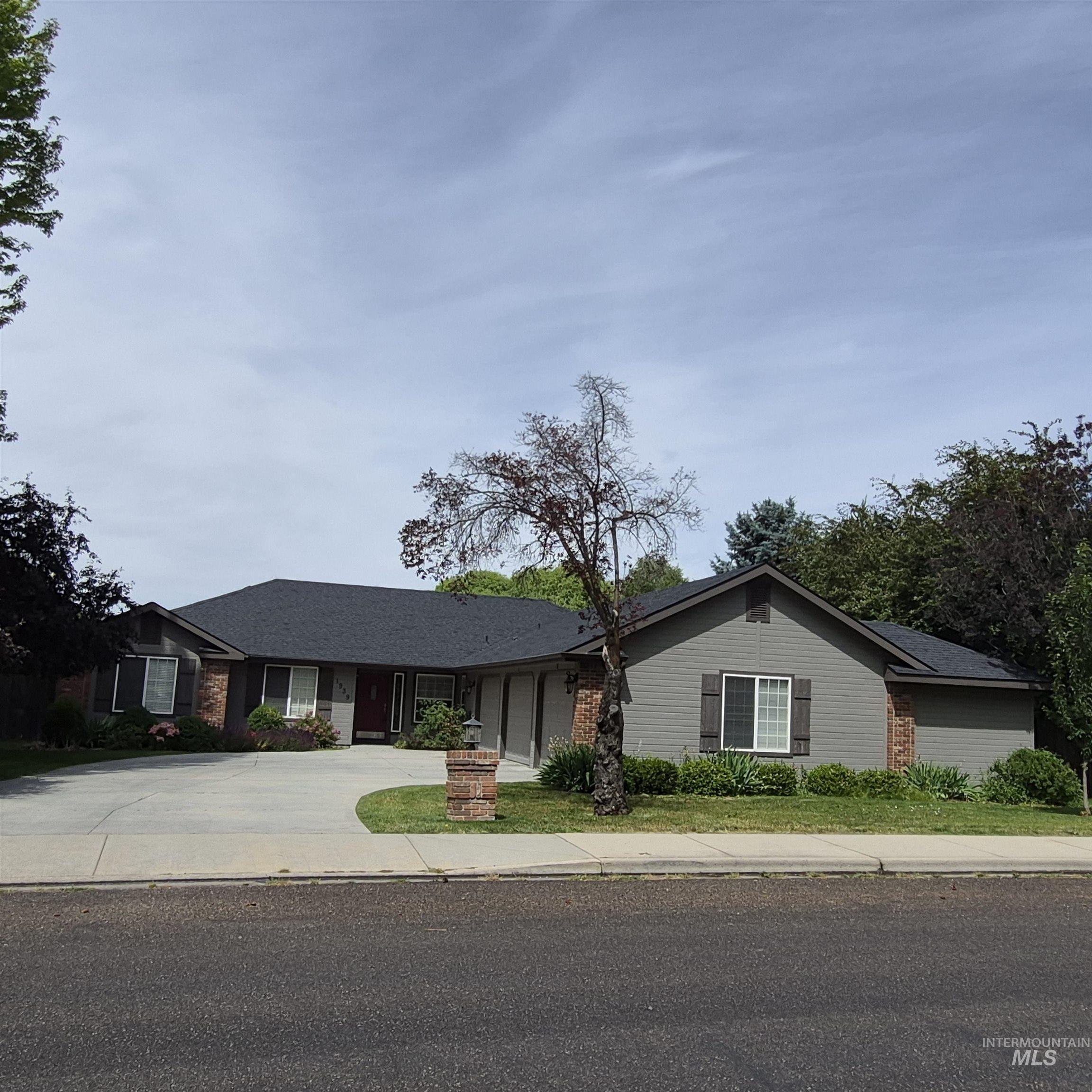 Contemporary ranch-style residence featuring a gray siding and brick facade, dark shingle roofing, a concrete driveway, and established landscaping