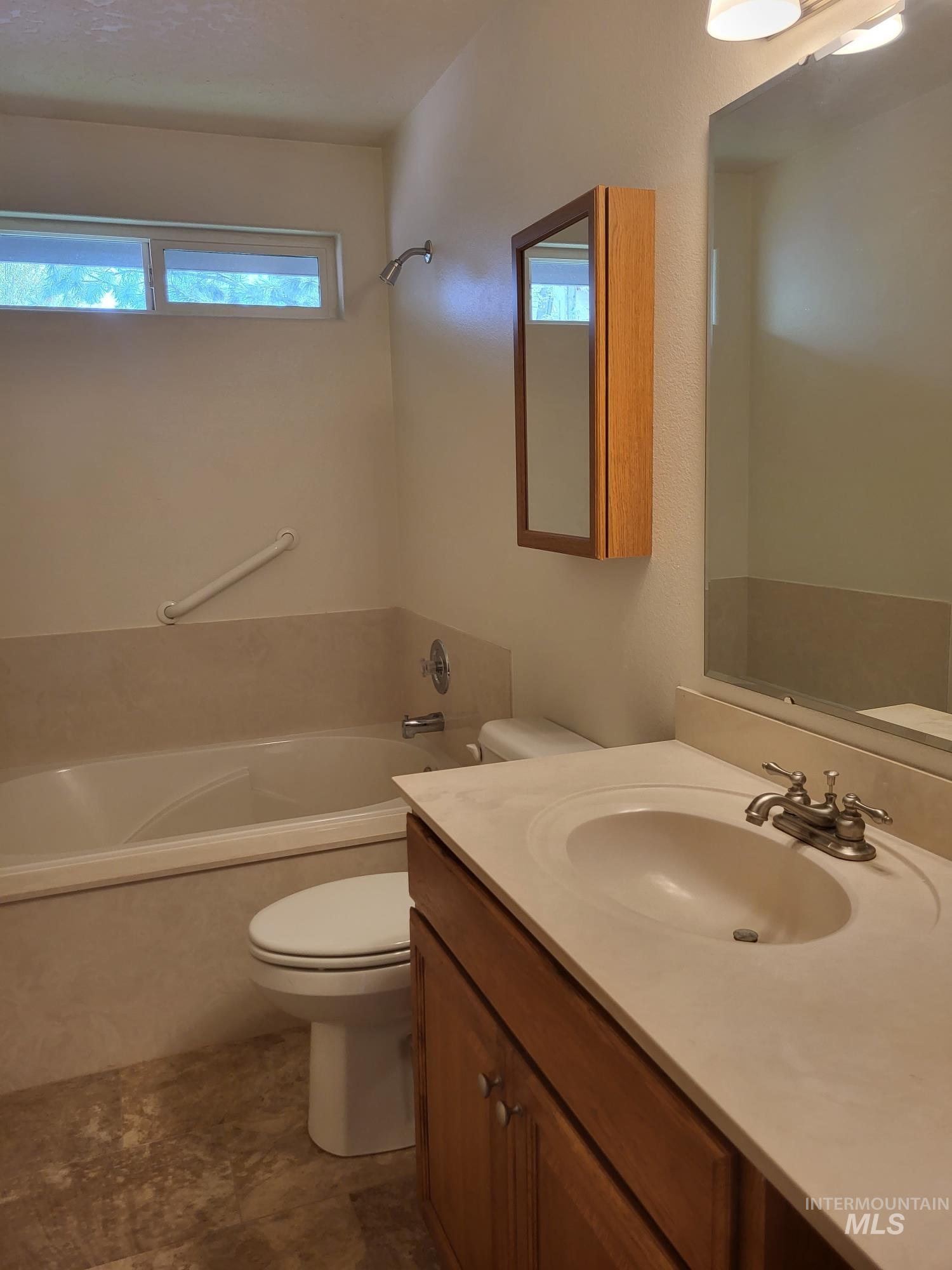 Bathroom featuring a light wood-finish vanity with an integrated sink, a large wall-mounted mirror, a built-in tub with grab bar, a window for natural light, and varied-tone flooring