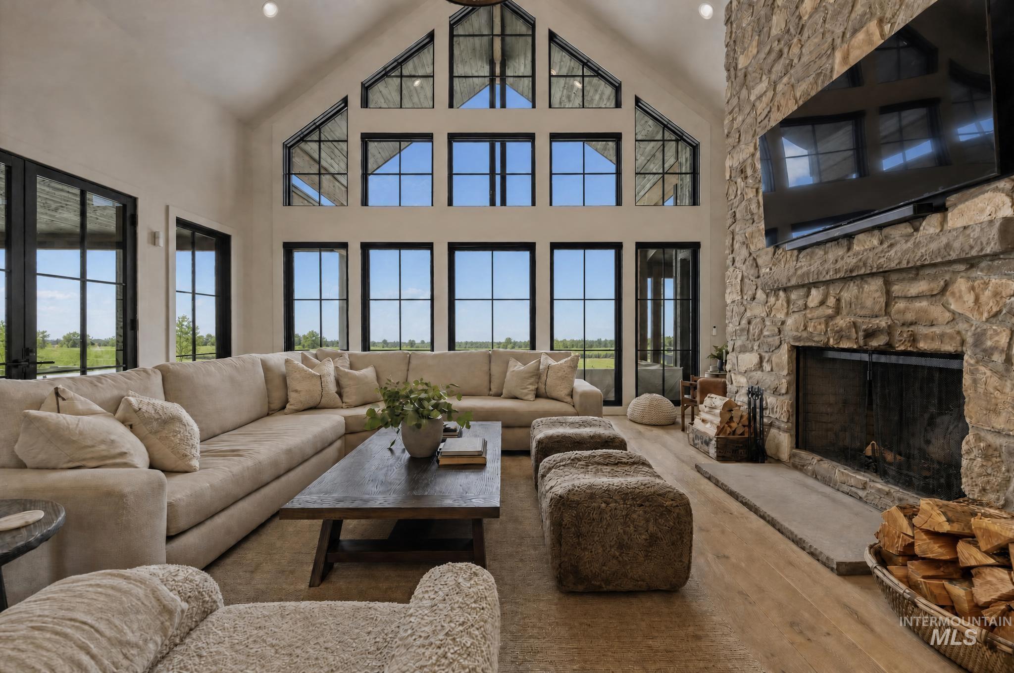 Spacious living area featuring a vaulted ceiling with exposed beams, an expansive stone fireplace, a wall of black-framed windows, wood-finish flooring, and recessed lighting