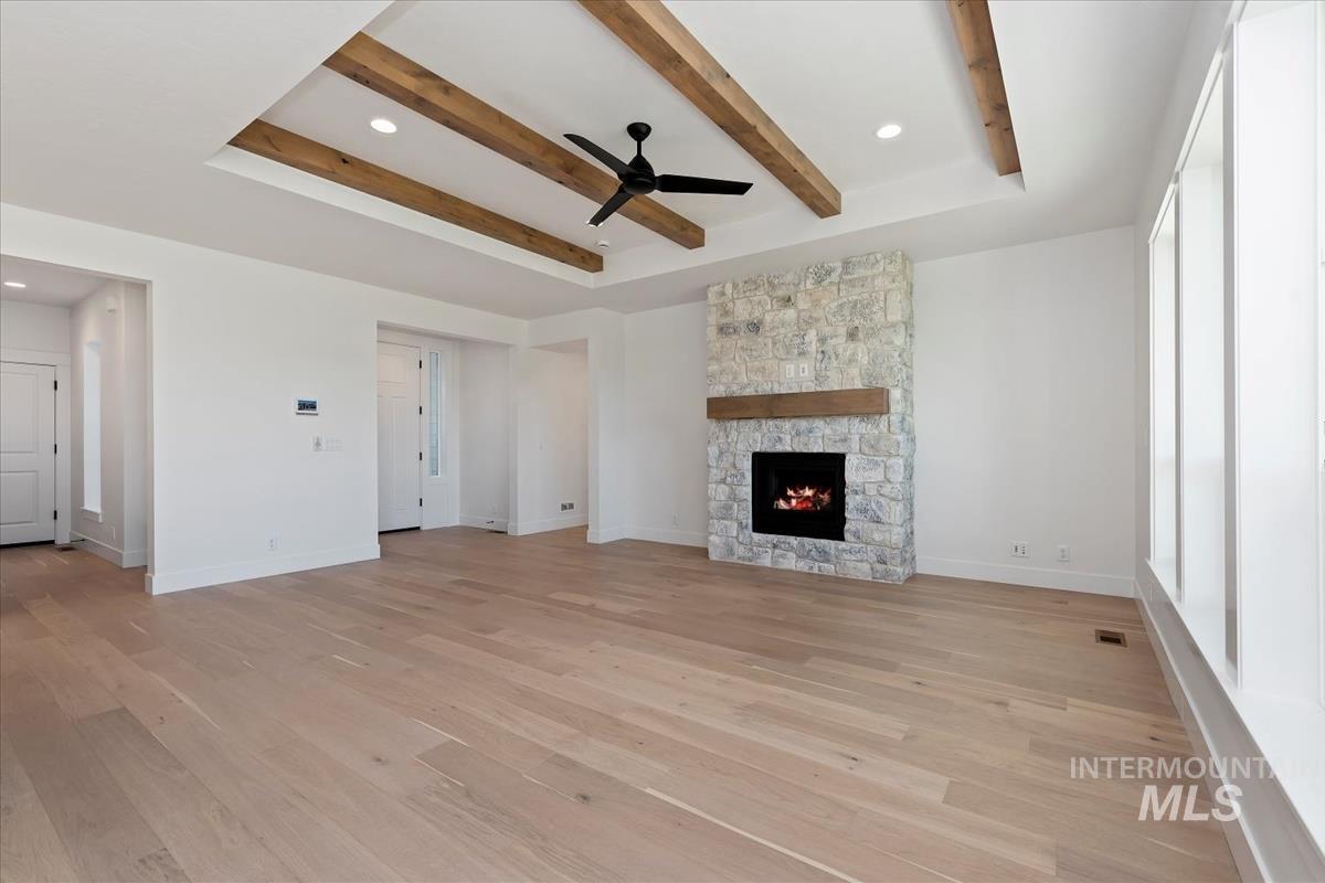 Spacious room featuring light wood-finish flooring, a stone-clad fireplace with a wood mantle, and a tray ceiling with exposed wood beams and a ceiling fan
