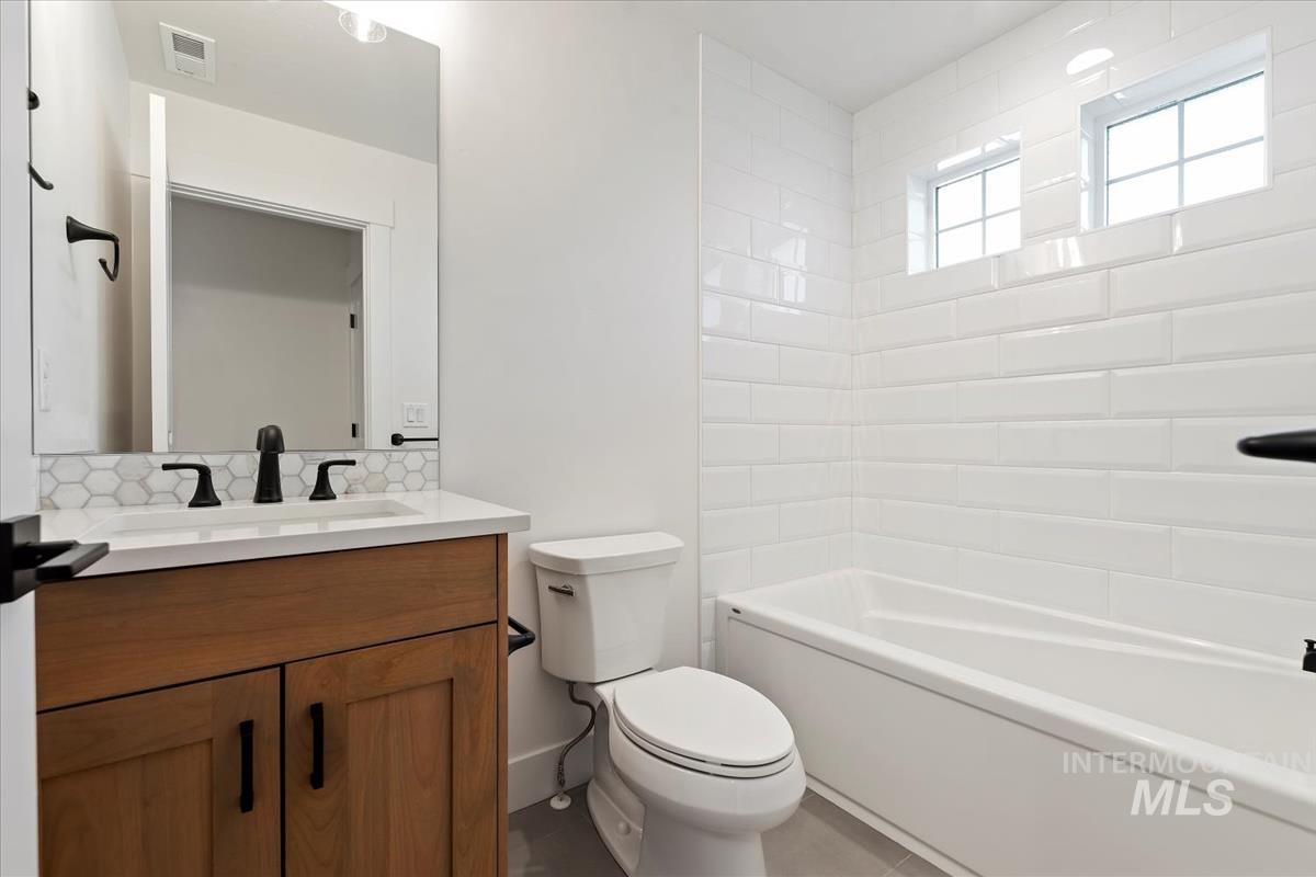 Bathroom featuring a wood-finish vanity with a white countertop, hexagonal tile backsplash, matte black fixtures, and a white subway tile tub surround with two transom windows