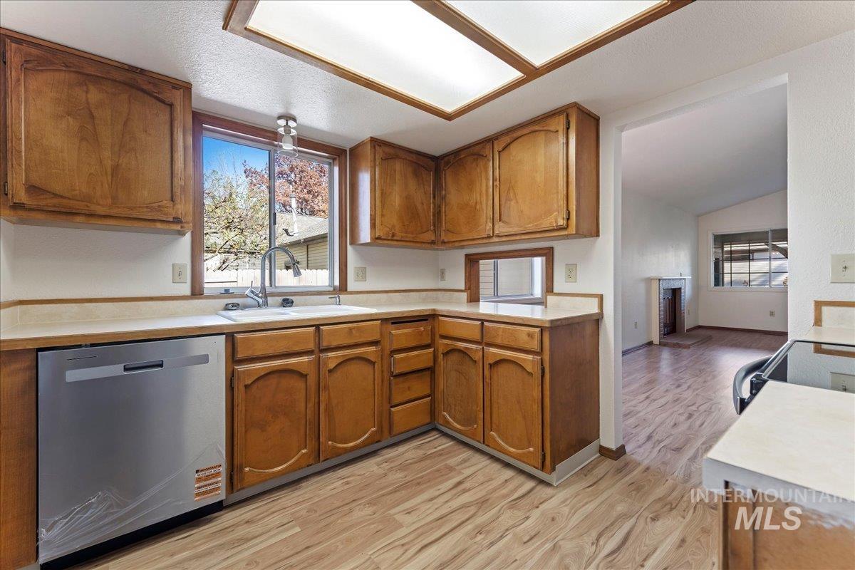 Kitchen featuring light countertops, brown cabinetry, vaulted ceiling, dishwasher, and light wood-style floors