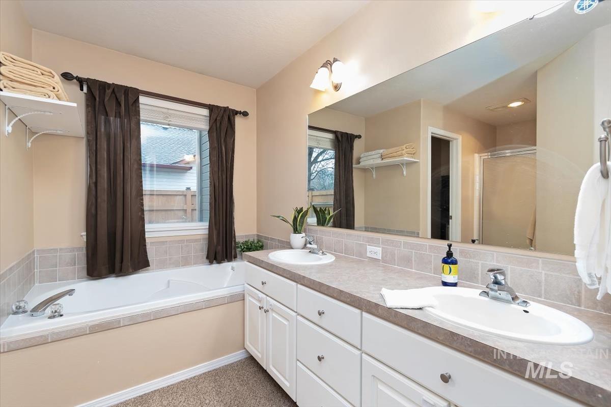 Bathroom featuring a dual vanity with white cabinetry, an integrated soaking tub, a large wall-mounted mirror, and tile backsplash
