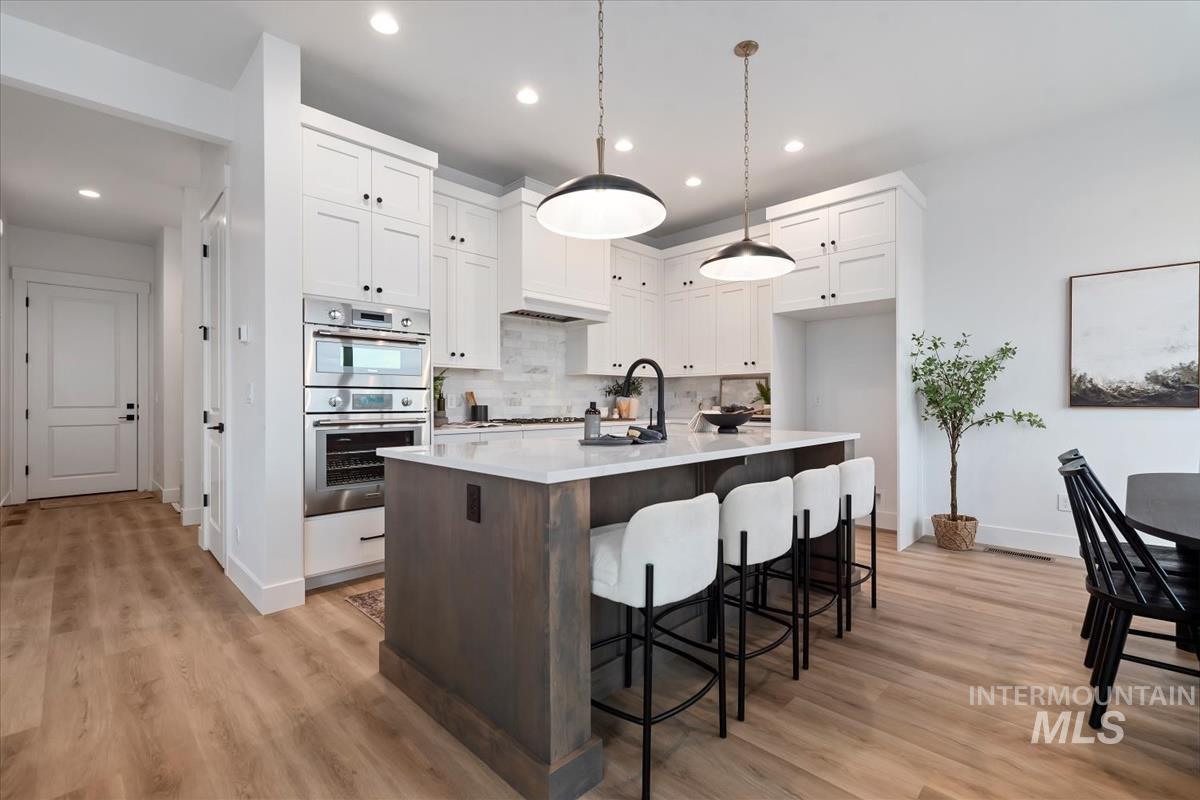 Kitchen featuring a center island with sink, white cabinets, hanging light fixtures, stainless steel double oven, and a kitchen bar