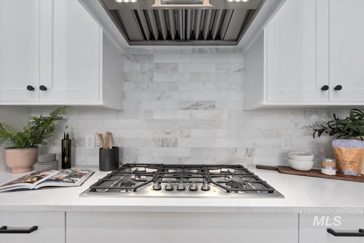 Kitchen featuring range hood, stainless steel gas stovetop, white cabinetry, backsplash, and light stone counters