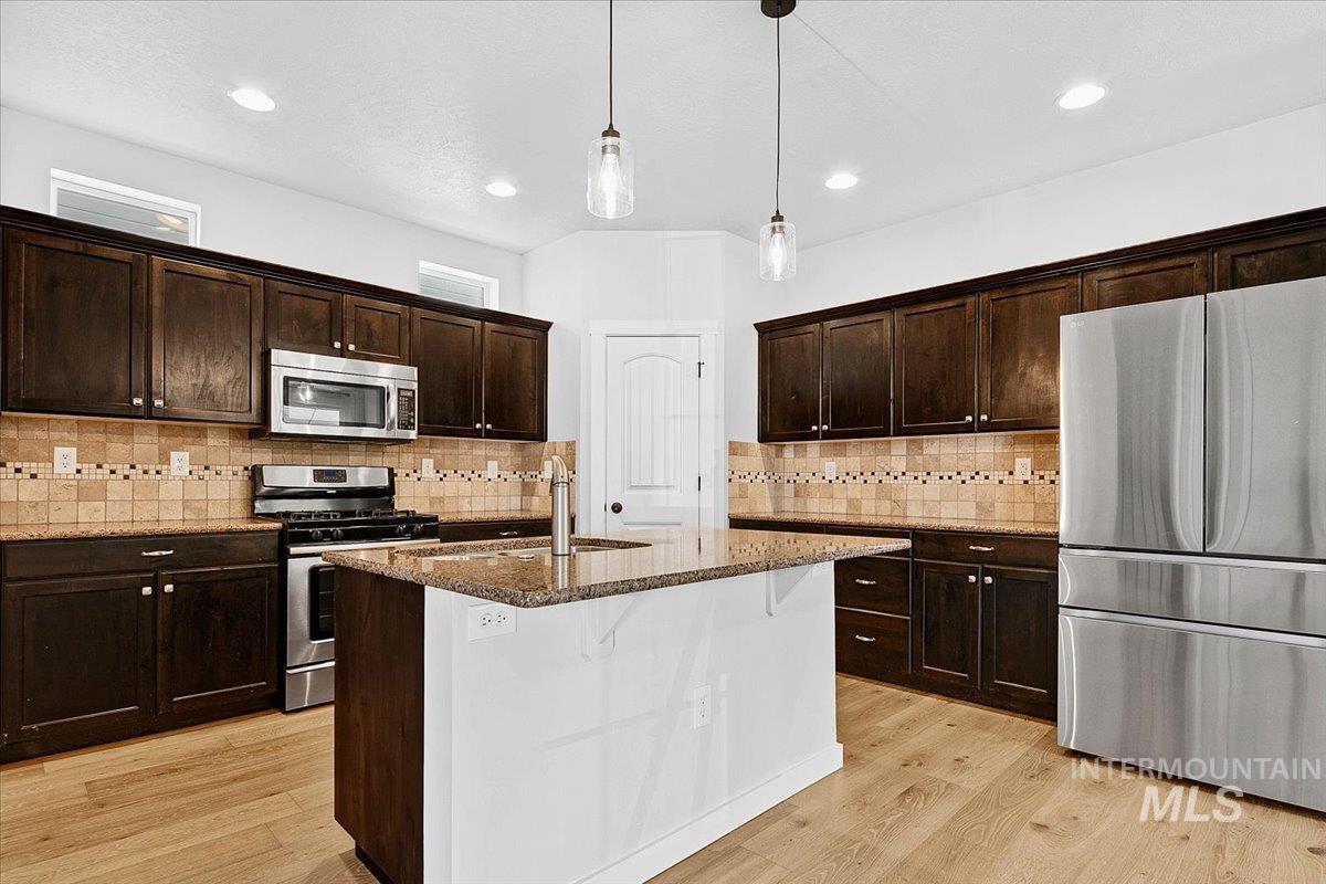 Kitchen featuring dark stone counters, stainless steel appliances, a center island with sink, light wood-style flooring, and dark wood finish cabinets