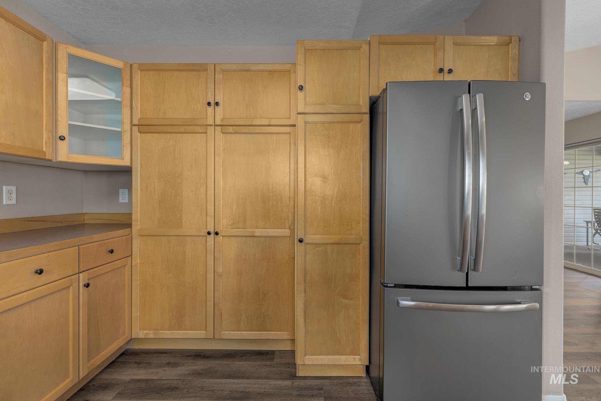 Kitchen with freestanding refrigerator, a textured ceiling, dark wood-style flooring, glass insert cabinets, and light brown cabinetry