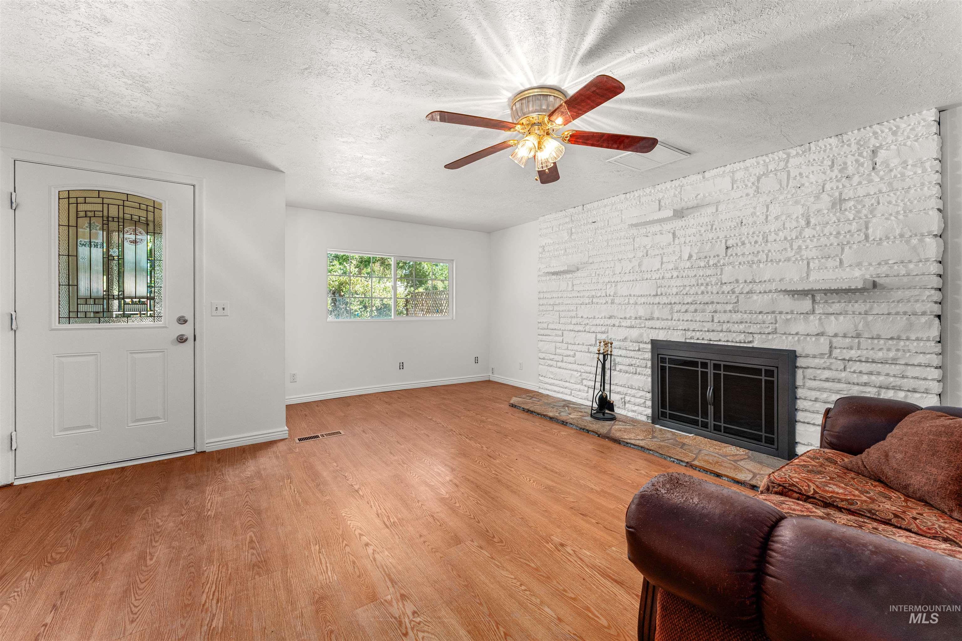 Living room with a stone fireplace, a textured ceiling, light wood finished floors, and a ceiling fan