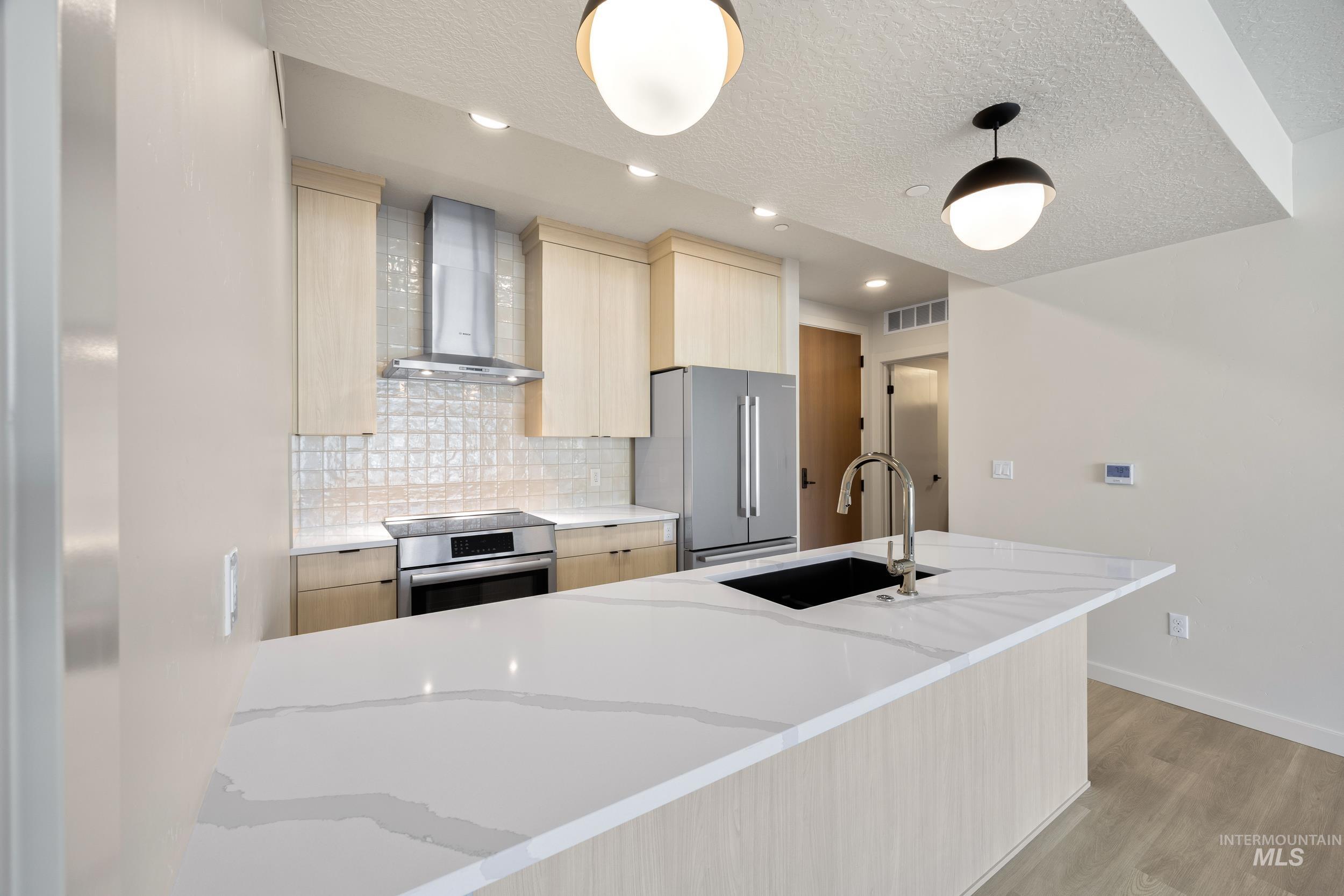 Kitchen featuring light brown cabinetry, tasteful backsplash, wall chimney range hood, light wood-style floors, and a textured ceiling