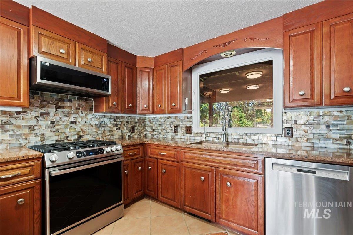 Kitchen featuring appliances with stainless steel finishes, brown cabinets, light tile patterned floors, light stone countertops, and backsplash