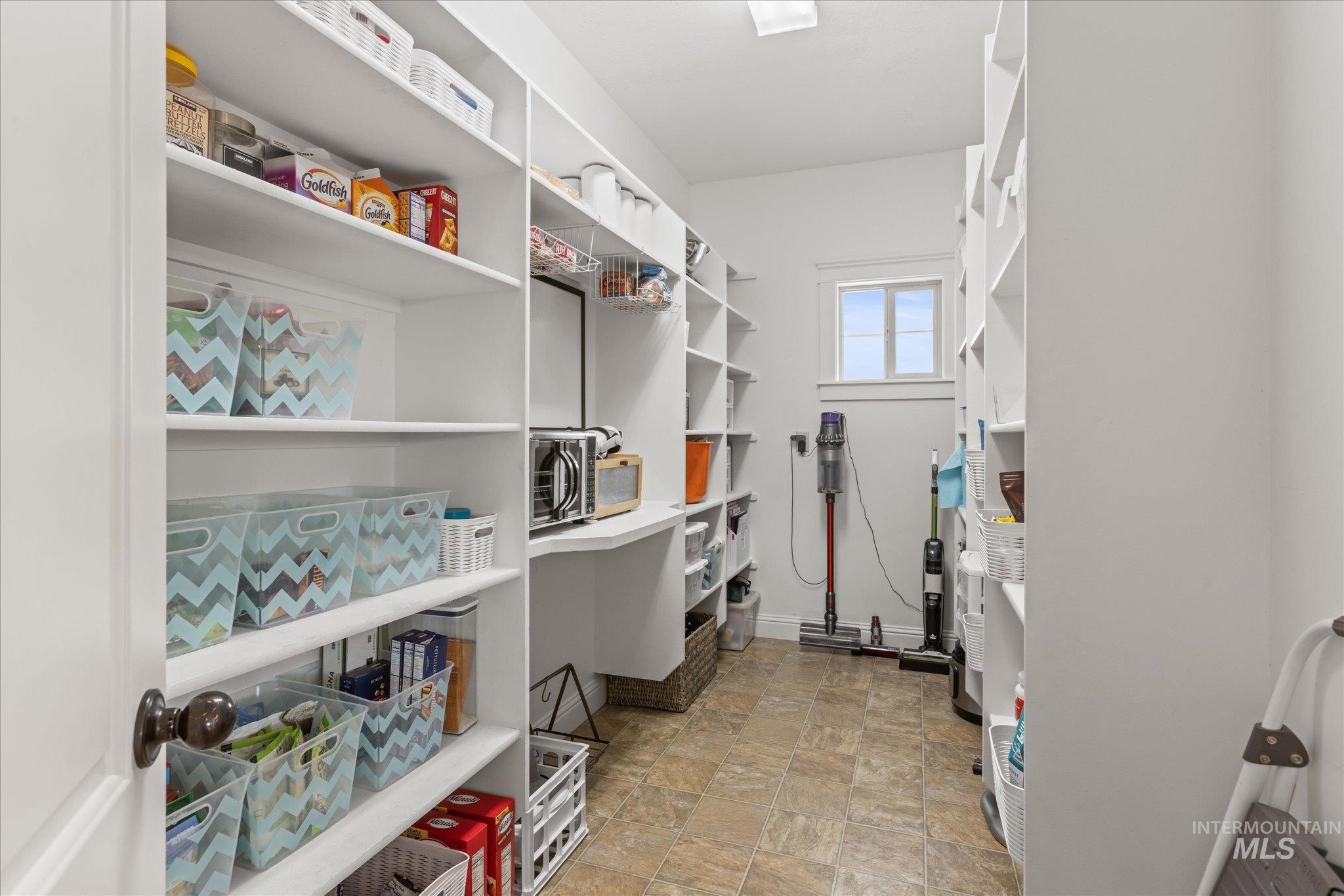 Spacious walk-in pantry featuring extensive white shelving, a built-in counter surface, tiled flooring, and a single window for natural light