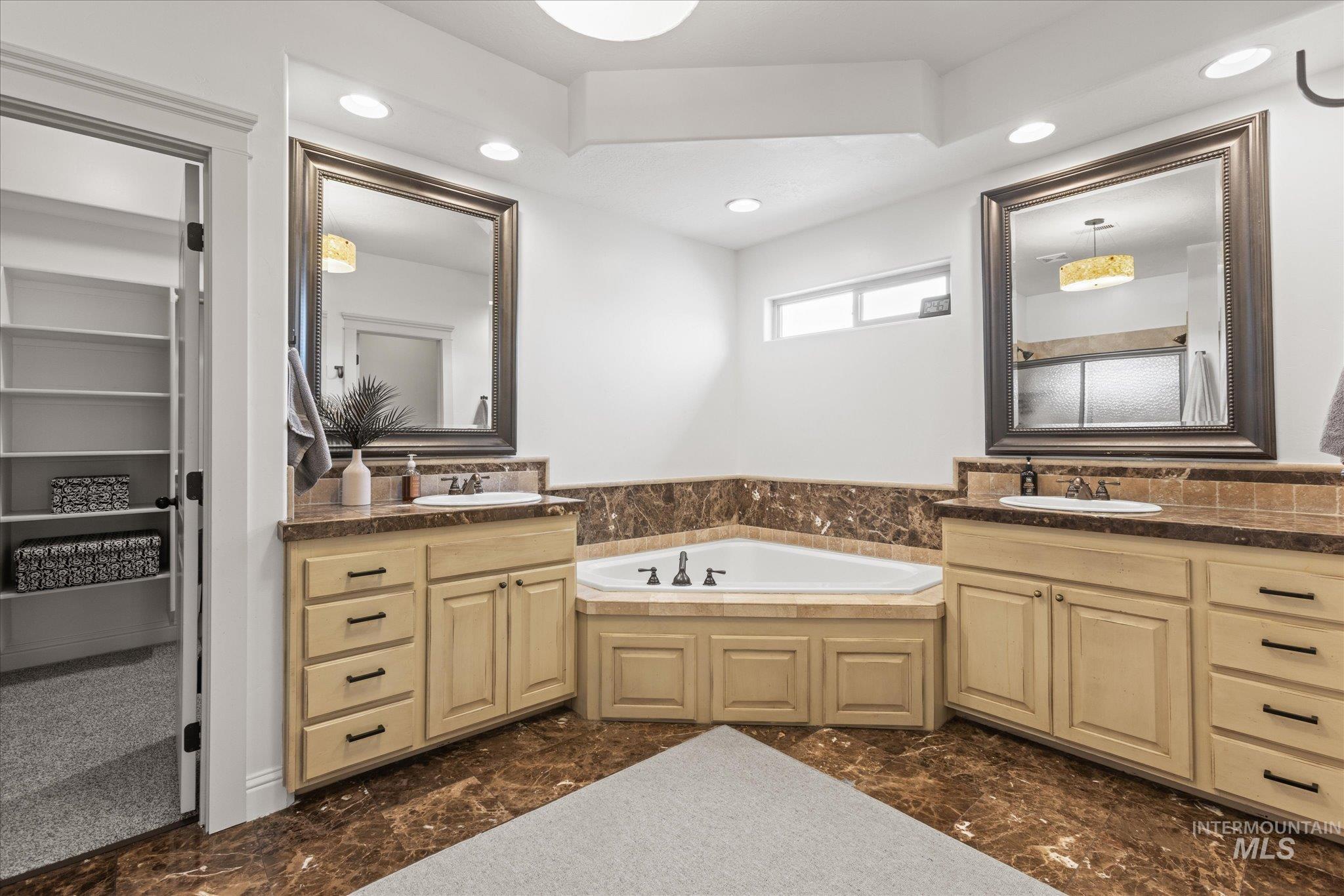 Dual vanity bathroom featuring a corner soaking tub, rich brown marble-finish flooring and wainscoting, light wood-finish cabinetry, and recessed lighting