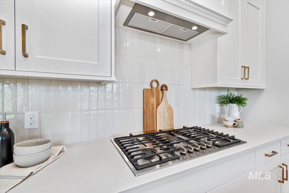 Kitchen view of white cabinets, custom range hood, stainless steel gas cooktop, and backsplash