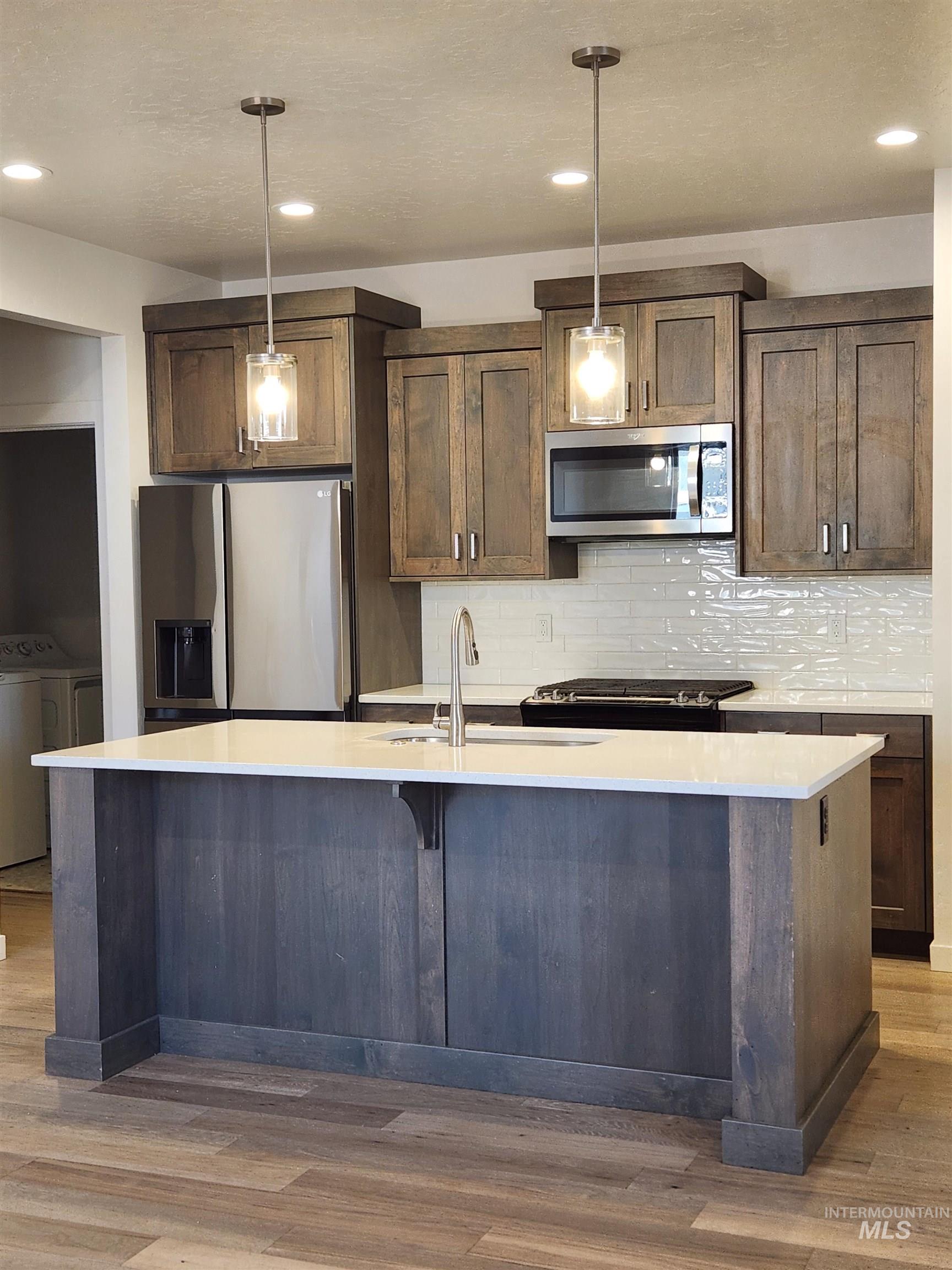Kitchen with stainless steel appliances, decorative light fixtures, an island with sink, light wood-style floors, and light stone countertops