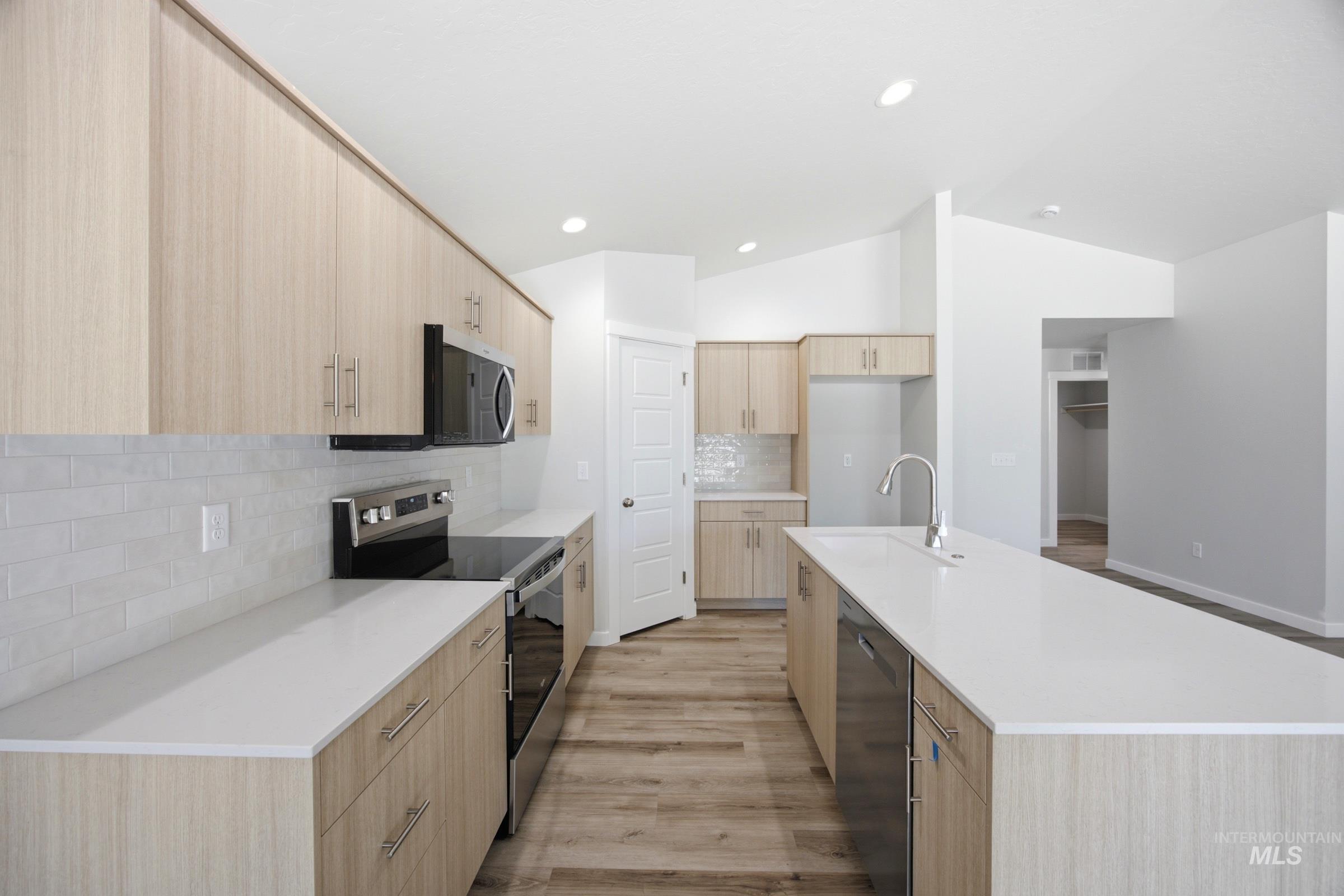 Open-concept kitchen featuring light wood-finish cabinetry, stainless steel appliances, white countertops, and a subway tile backsplash