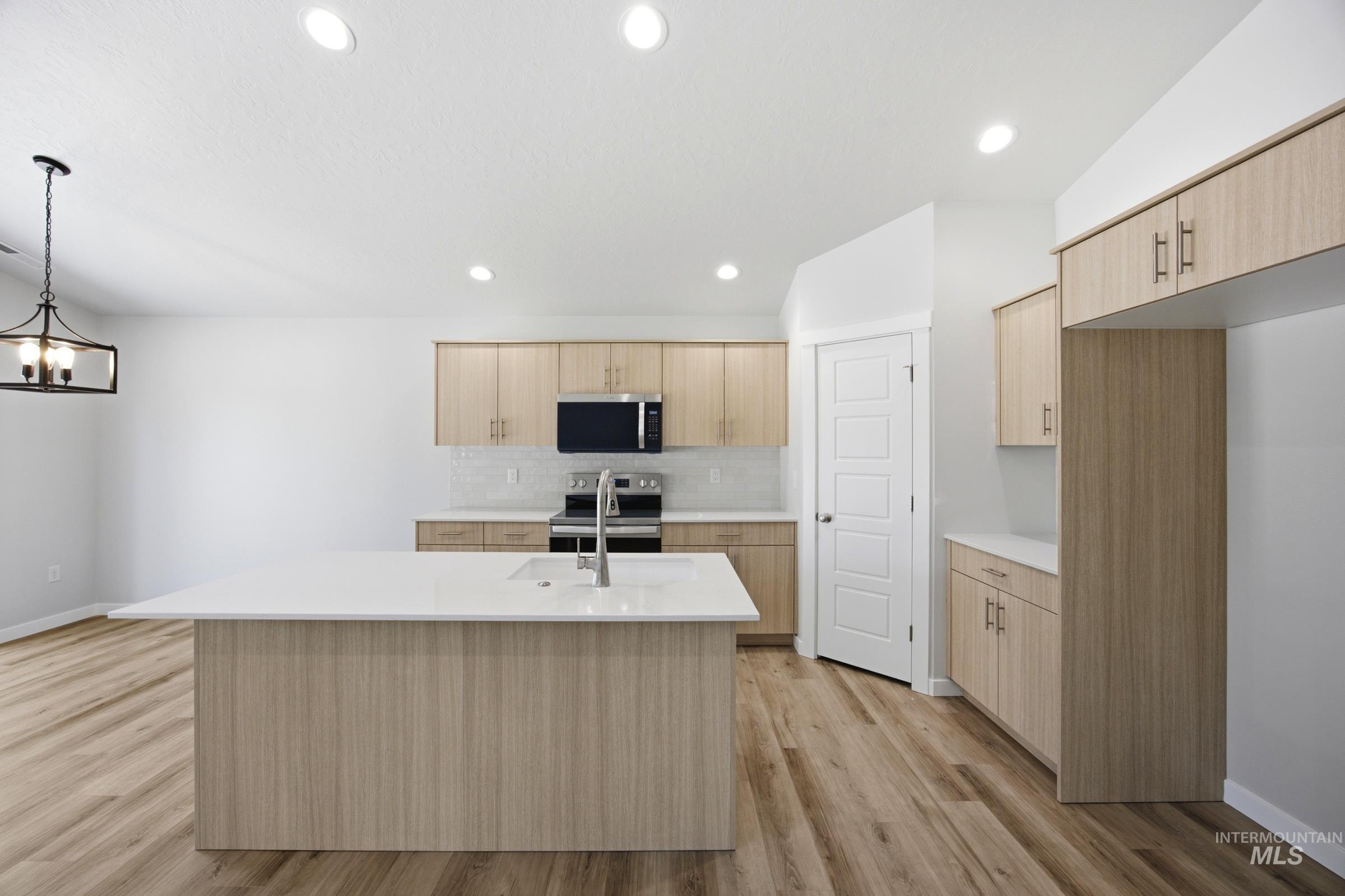 Open-concept kitchen featuring light wood-finish cabinetry, a large central island with an undermount sink, stainless steel appliances, and wood-finish flooring
