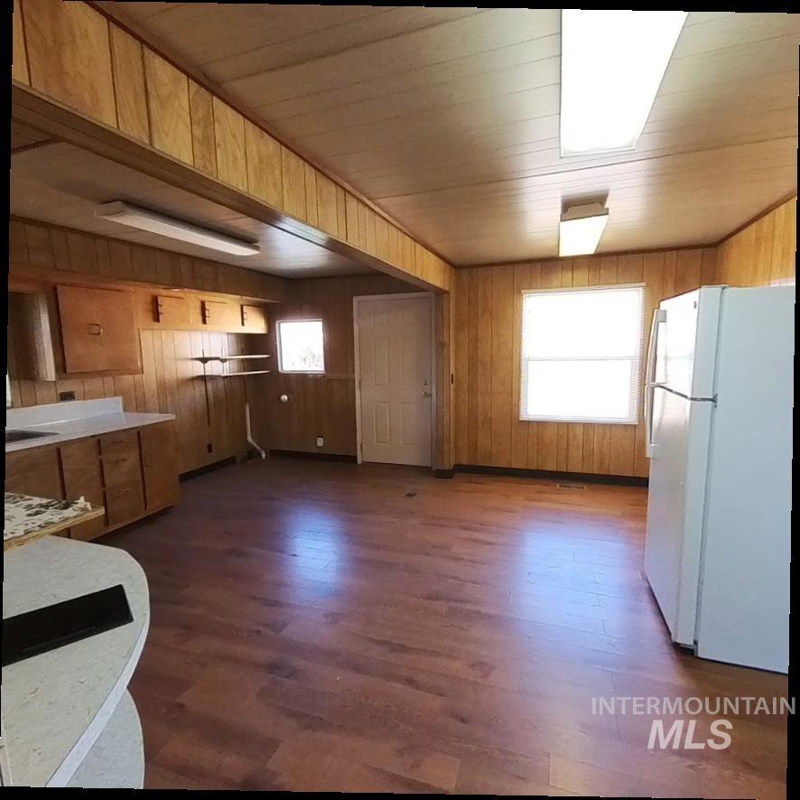 Kitchen with freestanding refrigerator, wooden walls, dark wood-type flooring, brown cabinetry, and light countertops