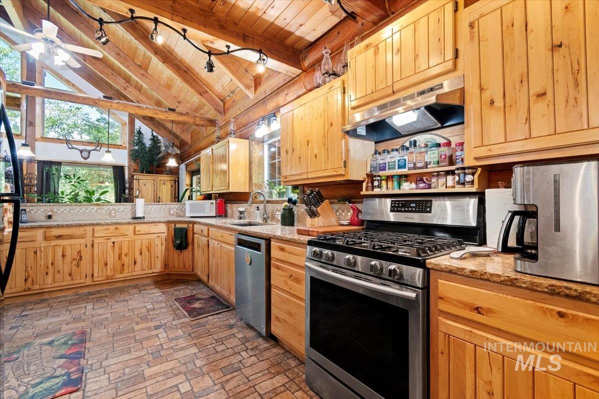 Kitchen with a high wooden beamed ceiling, stainless steel appliances, light stone counters, a ceiling fan, and light wood finish cabinets
