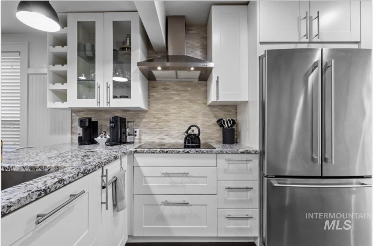 Kitchen with high end fridge, wall chimney exhaust hood, glass insert cabinets, light stone counters, and white cabinets