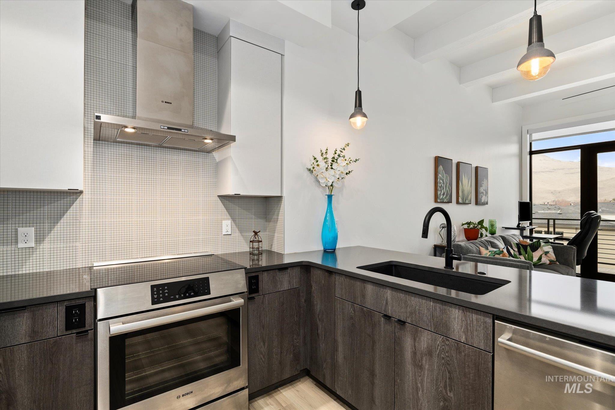 Kitchen featuring wall chimney range hood, stainless steel appliances, decorative backsplash, pendant lighting, and beamed ceiling