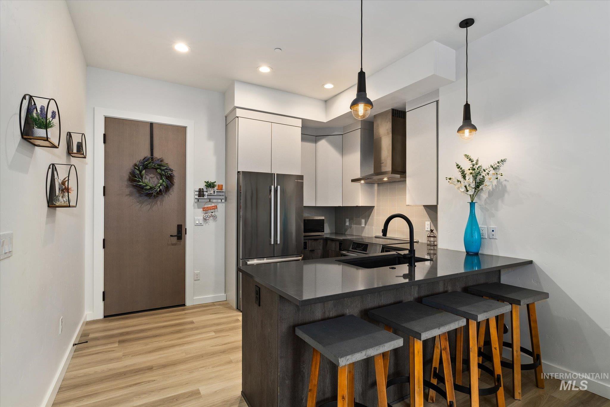 Kitchen featuring a kitchen bar, wall chimney range hood, hanging light fixtures, a peninsula, and backsplash