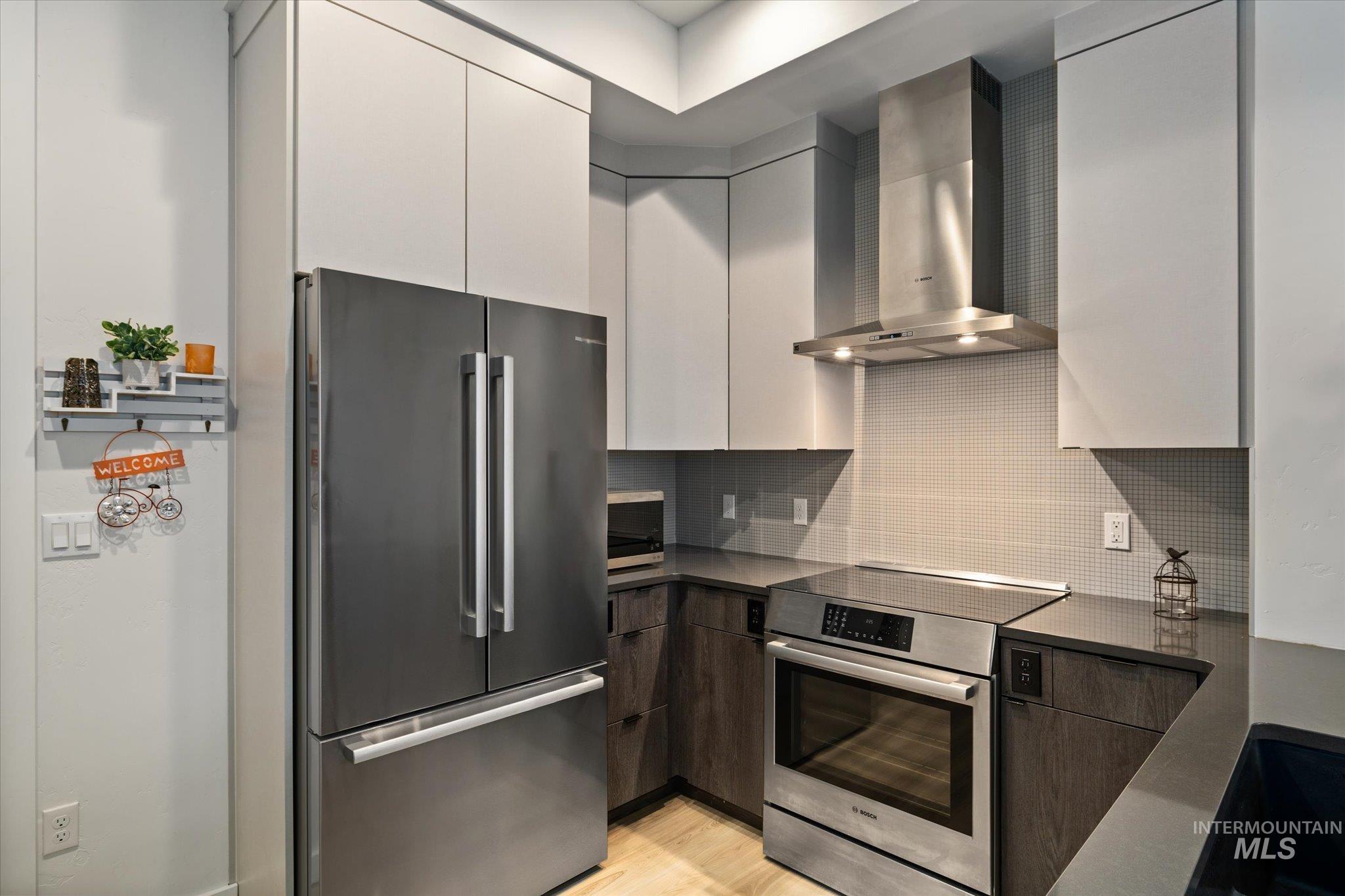 Kitchen with modern cabinets, appliances with stainless steel finishes, wall chimney range hood, backsplash, and light wood finished floors