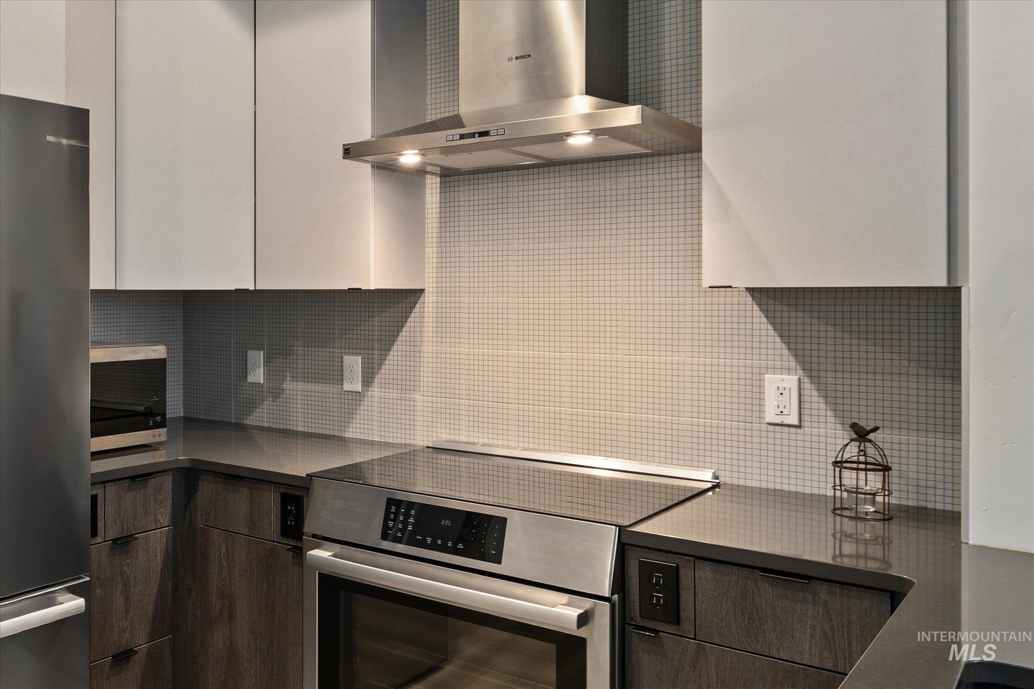 Kitchen featuring wall chimney exhaust hood, dark countertops, appliances with stainless steel finishes, decorative backsplash, and modern cabinets